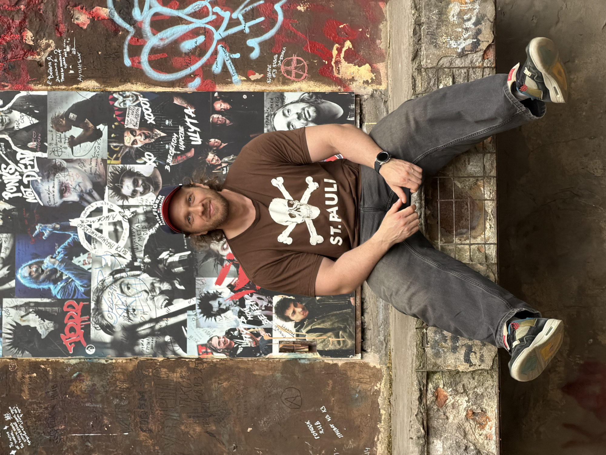 A man is sitting on a concrete ledge in front of a wall covered with posters and graffiti. The man is wearing a brown t-shirt with a skull and crossbones design and the words "ST. PAULI" below it, dark wash jeans, a baseball cap, and sneakers. He is looking at the camera with a slight smile. The wall behind him features a collage of black and white photographs of musicians and punk rock imagery, along with various graffiti tags and writings in Russian. The overall environment appears to be an urban outdoor setting, possibly an alley or a derelict building, with exposed brick and rubble visible on the ledge and surrounding area. The posters and graffiti suggest a connection to punk culture. Based on the Russian text visible in the graffiti and on some of the posters (e.g., "Понки не умрут" which translates to "Punks Not Dead"), the location is identified as Saint Petersburg, Russia.