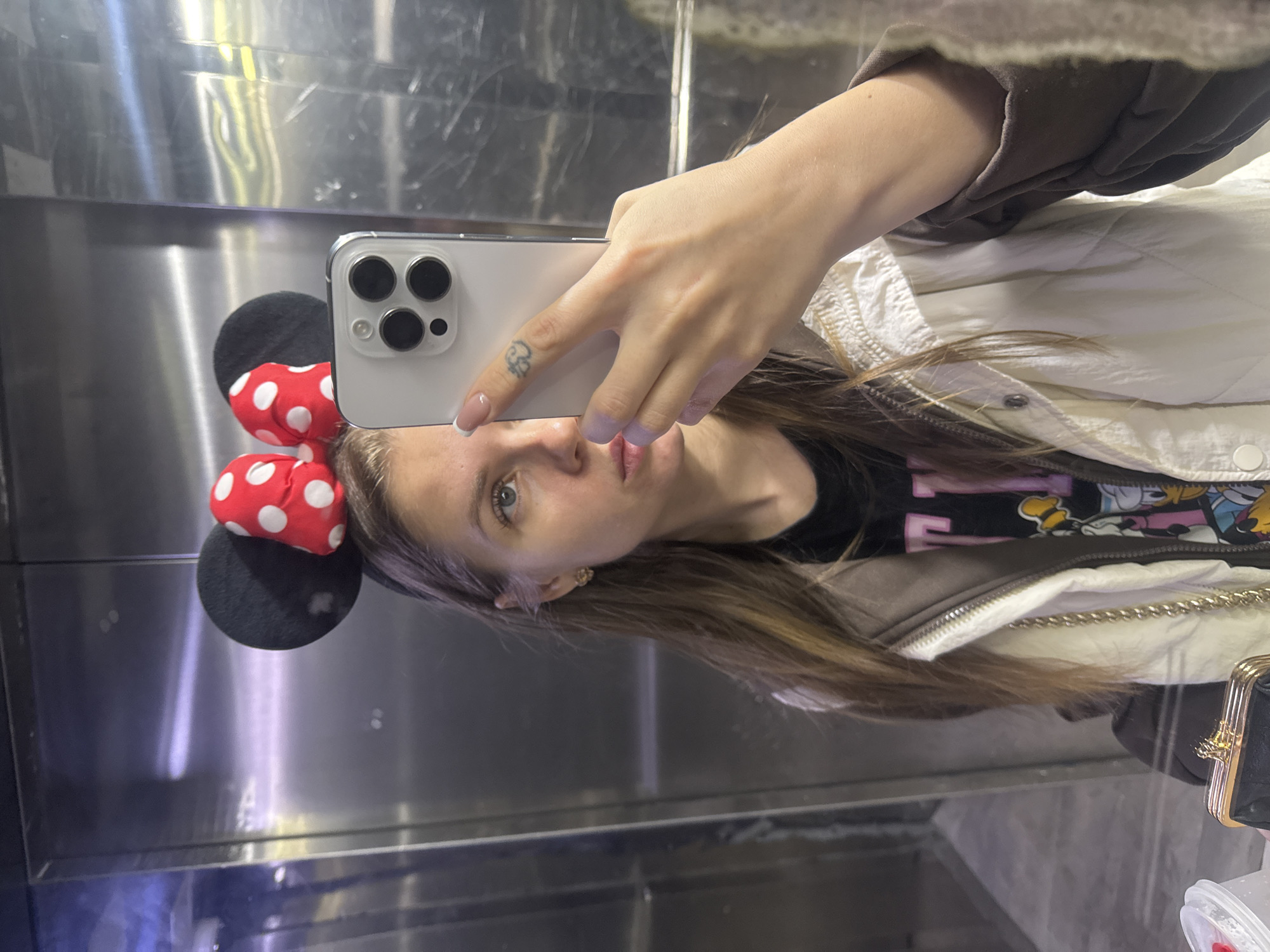 A female subject is depicted in a selfie taken within a brushed metallic elevator interior in Batumi, Georgia. The exact location within the city cannot be further specified.
The subject is a young adult female with light brown, long hair, light-colored eyes, and a fair complexion. She wears a black headband with black Mickey Mouse-style ears and a red, white-polka-dotted bow. Her right hand, featuring a small stylized cloud tattoo on the index finger and French-tipped fingernails, holds a silver-colored smartphone (identified as an iPhone 14 Pro or similar model due to its triple camera array) to capture the image, partially obscuring her face from the nose upwards on the left side. She is wearing a black graphic t-shirt, visible beneath an open two-tone jacket with a quilted cream-colored body and brown sleeves. A small gold-colored earring is present in her visible right ear. In the lower-left foreground, a black purse or clutch with a gold clasp is partially visible, alongside a clear plastic cup with a red lid and a white straw.
The elevator interior features brushed stainless steel or similar metallic panels, displaying reflections of overhead lighting. The floor appears dark and slightly reflective.