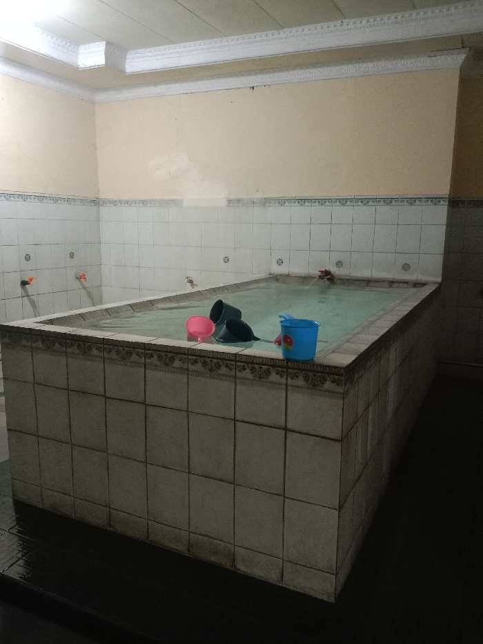 The image shows an indoor tiled rectangular pool filled with water and steam, likely in a public bathhouse located at latitude -6.976, longitude 110.331 in Tugu, Indonesia. The pool is surrounded by white tiles with a decorative border. There are four objects visible in the pool: a pink plastic bucket, a blue plastic bucket, and two dark blue plastic buckets. Two faucets are visible on the wall, and a third faucet appears to be protruding from the pool at the far end. The lighting is dim, creating a hazy atmosphere. The walls are tiled in white and have a decorative trim along the top. There are no people visible in the image. The floor surrounding the pool is dark and appears to be tiled. The image captures a simple, functional space likely used for communal bathing.