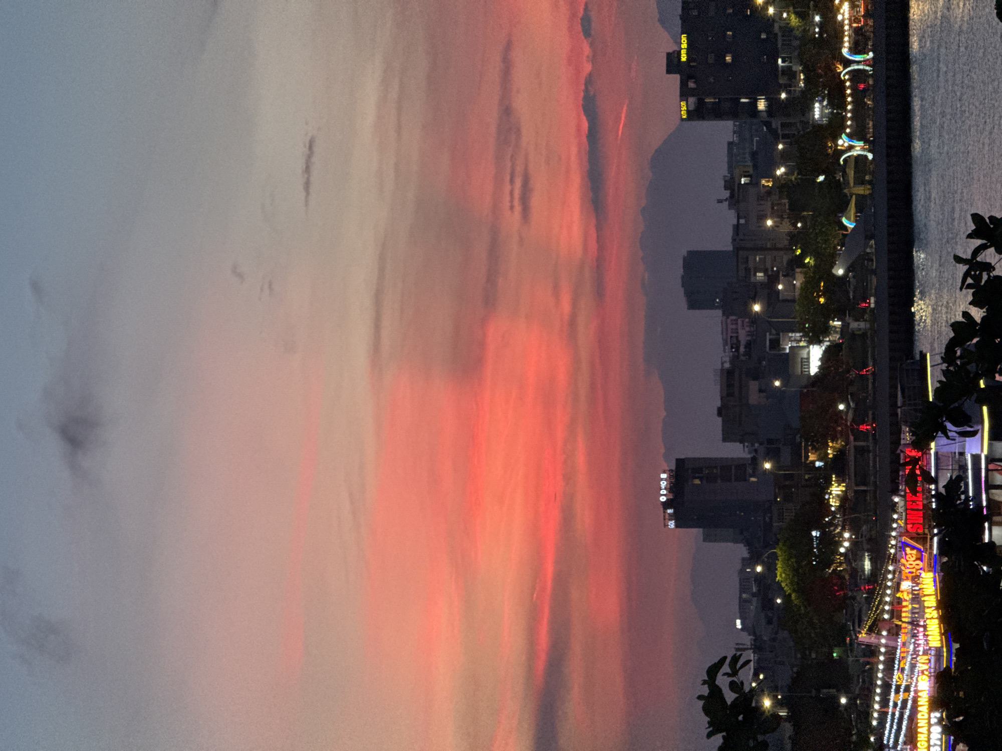 The image shows a vibrant sunset over the Han River in Da Nang, Vietnam. The sky is a dramatic display of pink, orange, and purple hues, with scattered dark clouds. In the foreground, the dark silhouettes of trees frame the scene. The middle ground features the Da Nang cityscape, with various buildings and lights illuminated against the twilight sky. Prominent buildings include the "GOLDEN HOTEL" and "KIM SON" buildings. Along the riverfront, there are numerous brightly lit boats and structures, suggesting a lively evening atmosphere. The Han River itself is visible, reflecting the lights of the city and the colors of the sunset. In the distance, hazy mountains form a soft backdrop to the urban landscape. The overall impression is one of urban beauty at dusk, with natural light and artificial illumination creating a captivating scene.