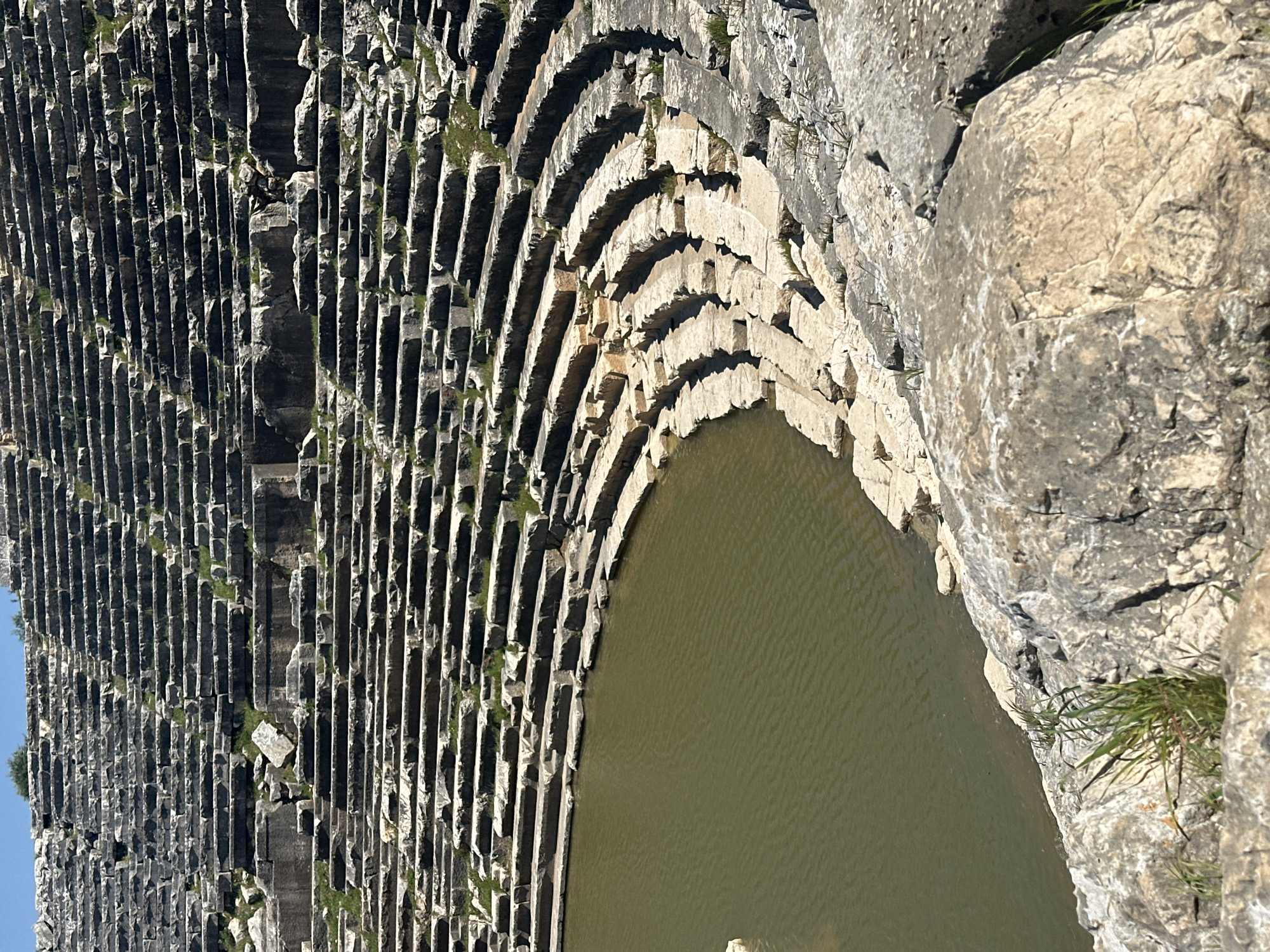 The image shows a partial view of the ancient theatre in Seydikemer, Turkey, with a focus on the seating rows and the orchestra area now filled with water. The seats are constructed of light-colored stone. Water fills the orchestra area. A large rock formation is visible on the right. Patches of green moss or small plants grow in between the stones. The image is shot from a high angle.