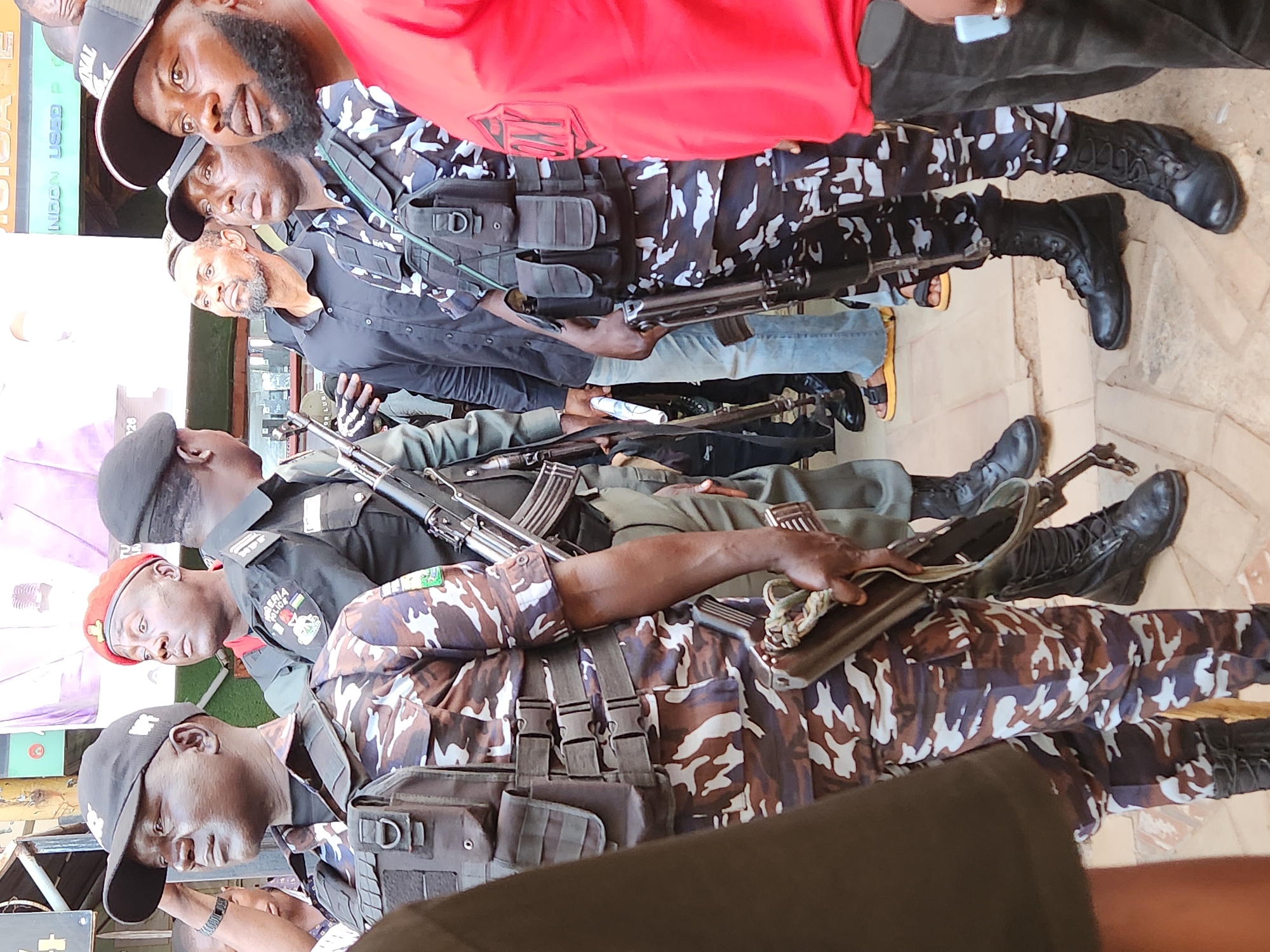 The image displays a group of men, several of whom are armed and dressed in various types of camouflage and official uniforms, in an outdoor setting during the daytime.
In the foreground and midground, at least five men are visible. One individual on the far left wears a dark military-style uniform and a red beret. Another man to his right wears a camouflage uniform featuring brown, dark green, and white patterns, a tactical vest, and black combat boots. He is holding a rifle, possibly an AK-pattern. Further right, a man in a black uniform with a "NIGERIA POLICE" patch and crest on his shoulder is visible, also wearing a black cap.
Towards the right side of the image, another man in a dark blue/black and white camouflage uniform, tactical vest, and black combat boots is holding a rifle, which appears to be a Heckler & Koch G3 or similar battle rifle. He stands next to a civilian in a black shirt and jeans, who is also wearing black combat boots. At the top of the frame, partially obscured, is a man in a red shirt and a black cap, looking towards the camera. Another man in a black cap is also visible near him.
The individuals appear to be standing or moving in an urban or market-like area with tiled or paved ground. In the background, an inverted and partially obscured sign is visible, with discernible text fragments such as "NDON USED P" and "SICA," possibly indicating a business like "LONDON USED PHONES". The bright lighting suggests it is daytime.