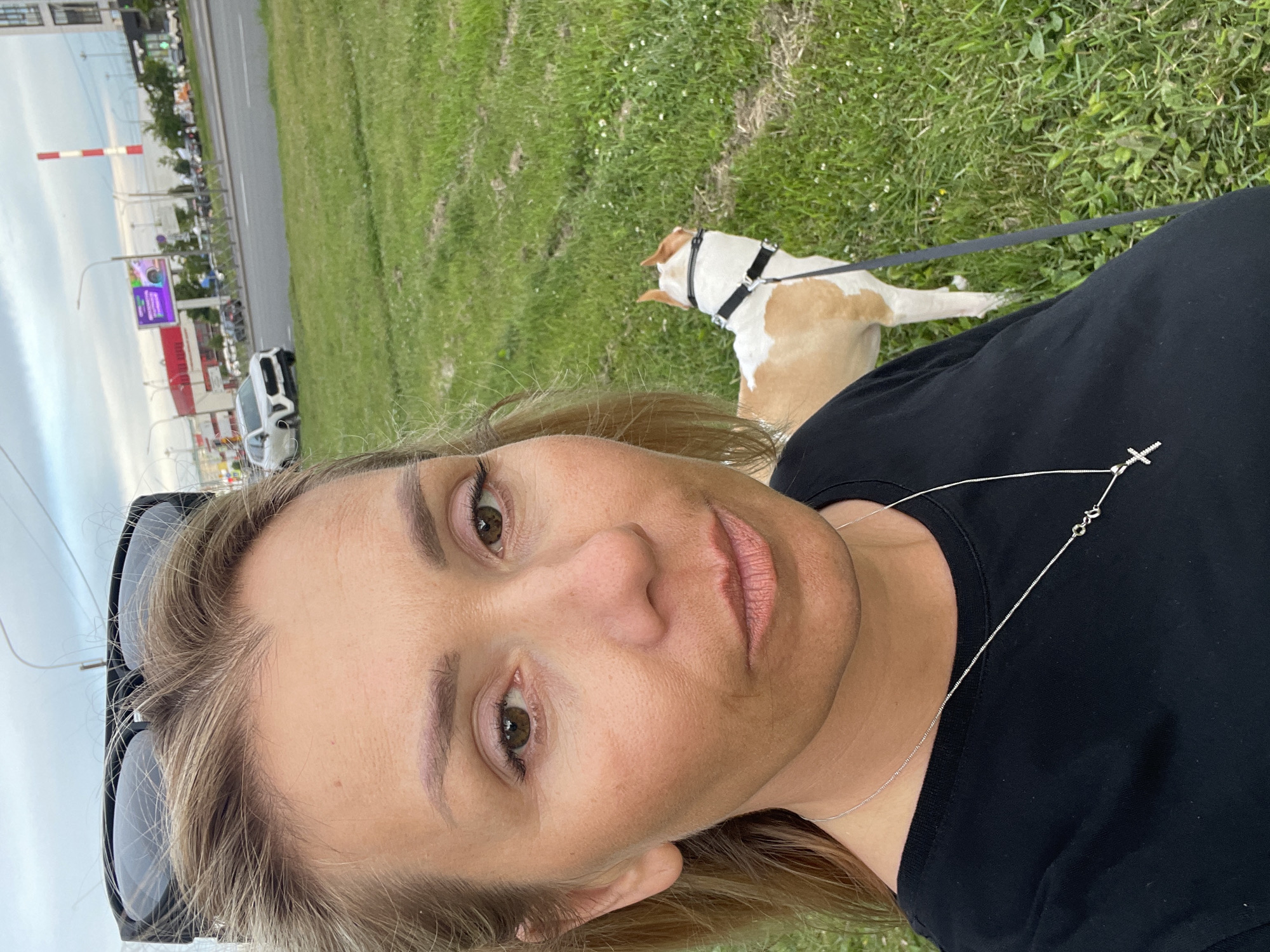 A medium shot shows a fair-skinned woman with blonde hair pulled back, wearing sunglasses perched on her head, and a black t-shirt with a silver cross necklace. She is standing outside in a grassy area. To her right, a tan and white dog on a leash stands facing away from the camera. In the background, a road with cars and buildings is visible under a cloudy sky. The location is identified as Unknown, Unknown.