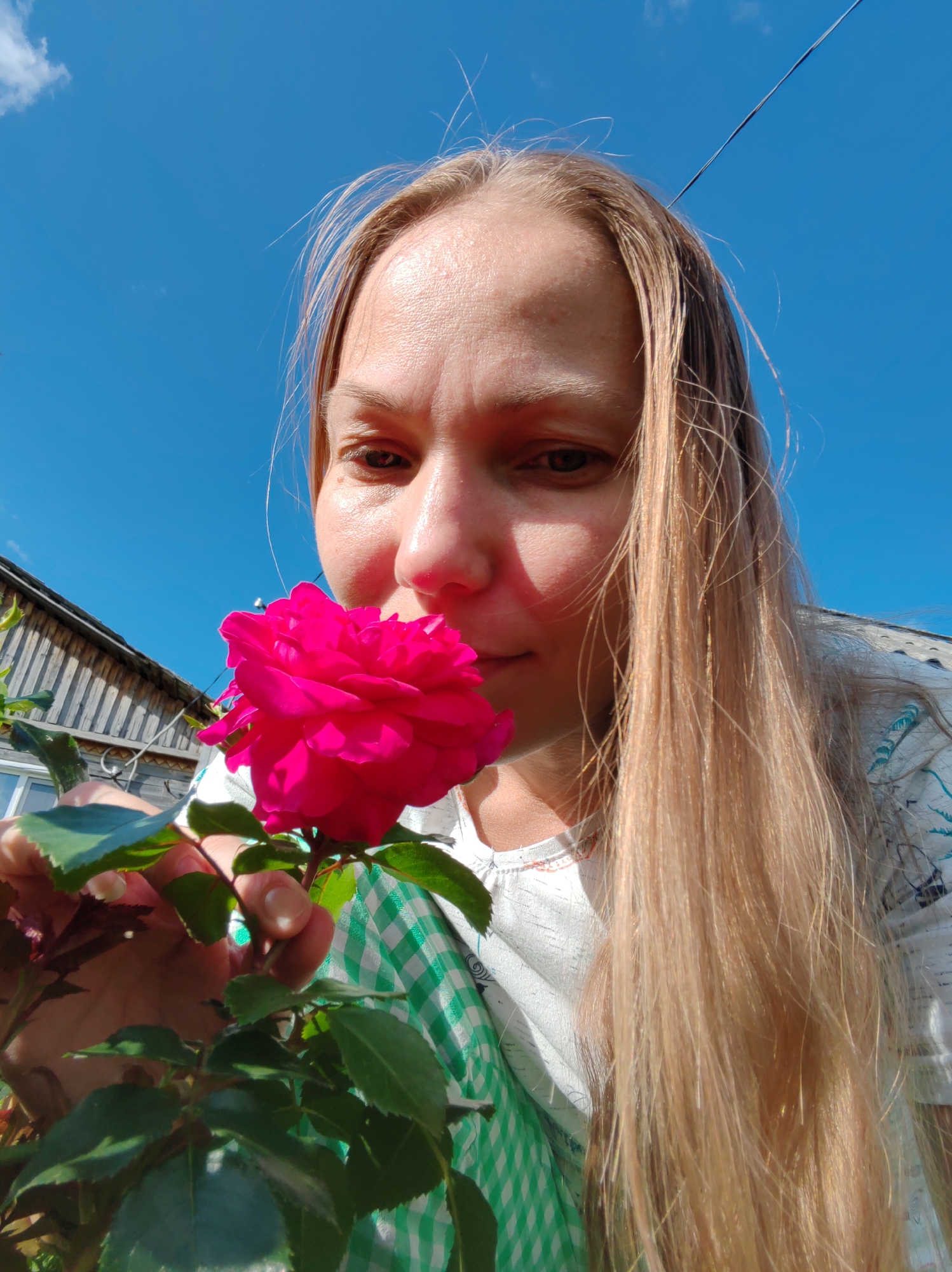 A close-up, low-angle shot shows a Caucasian woman with long blonde hair smelling a vibrant pink rose. She is outdoors, with a clear blue sky above. To the left, a portion of a wooden structure, possibly a house or shed, is visible. She is wearing a white shirt with a blue and white checkered fabric peeking out from underneath. The image appears to have been taken in Чухлэм, Russia, on a sunny day.