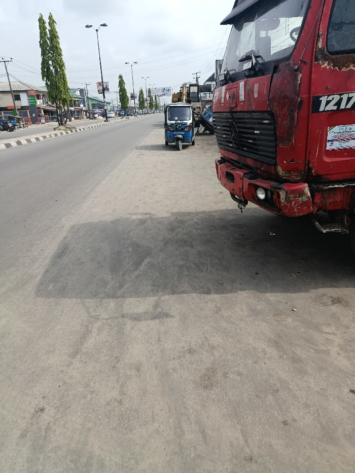 The image depicts a daytime urban street scene in Warri, Nigeria. In the immediate right foreground, the front portion of a heavily weathered and rust-damaged red heavy-duty truck is visible, displaying "1217" on its side and a Mercedes-Benz emblem on its grimy front grille. The front bumper is discolored, chipped, and appears to have a single functional headlight on the left, with a missing or damaged light on the right side.
Further down the dirt and concrete shoulder, slightly to the left of the red truck, a blue and white tricycle, commonly known as a Keke Napep, is parked. The word "VICTORY" is partially visible on its front.
To the left of these vehicles, a multi-lane asphalt road stretches into the background, separated by a median barrier painted with black and white stripes. Tall, slender green trees and black streetlights with double arms line the road and median. In the background on the left, a sequence of commercial buildings with various signs, including what appears to be a fuel station, flank the street.
The sky is overcast with visible clouds, indicating daylight conditions. Shadows from the vehicles and potential unseen structures fall across the shoulder and the edge of the road, suggesting illumination from the upper left. No distinct human figures or active interactions are clearly visible within the frame.