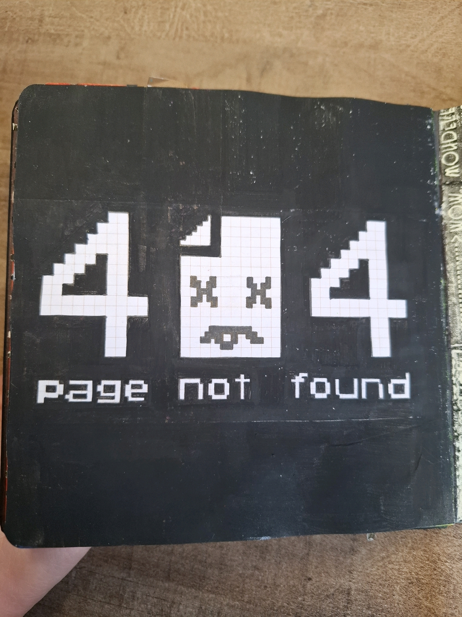 The image shows a close-up of a black notebook with a "404 Page Not Found" error message depicted in pixelated white text and graphics. The error message includes a stylized representation of a document with "X"s for eyes and a sad mouth, as well as two large "4"s. The text "page not found" is written below the graphic. The notebook is lying on a wooden surface, possibly a table. A hand is partially visible in the bottom left corner, suggesting someone is holding or presenting the notebook. The background is out of focus but appears to be a light-colored, textured surface. There is no discernible information or visual cues in the image that would indicate a location in Tyumen, Russia.