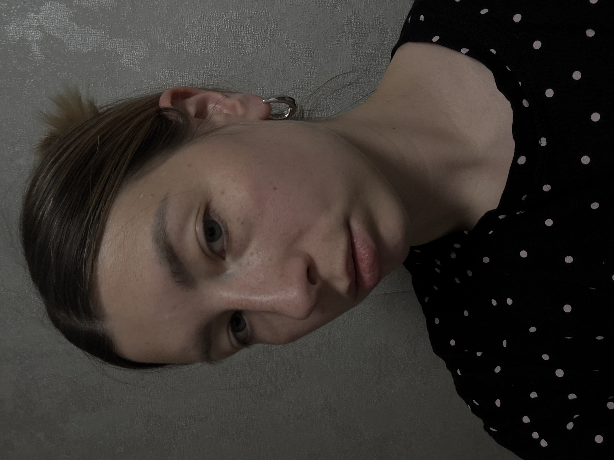 A person is facing the camera, turned slightly to the side. The person has fair skin, light brown hair pulled back, and is wearing a black top with white polka dots. A silver hoop earring is visible in the left ear. The background is a plain grey wall. The location is not identifiable as Moscow, Russia.