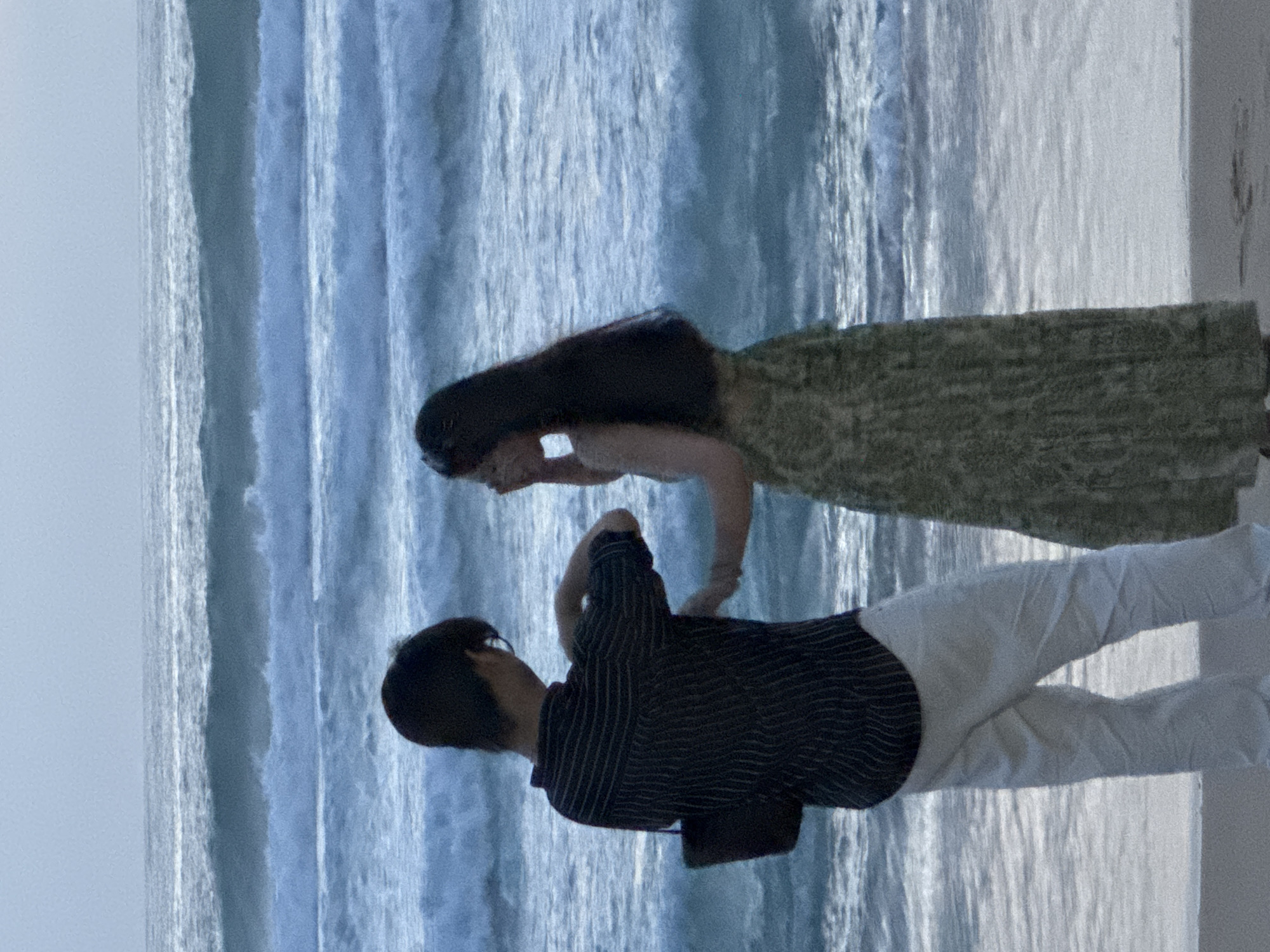 Two individuals, a male and a female, stand on a sandy beach with the ocean in the background. The male, facing away from the camera, wears a dark, pinstriped shirt and light-colored pants. He holds a dark bag in his left hand. The female, also facing away, wears a patterned dress. Her right hand is raised to her face. The ocean displays multiple rows of waves under a pale sky. The setting appears to be Karon, Thailand.