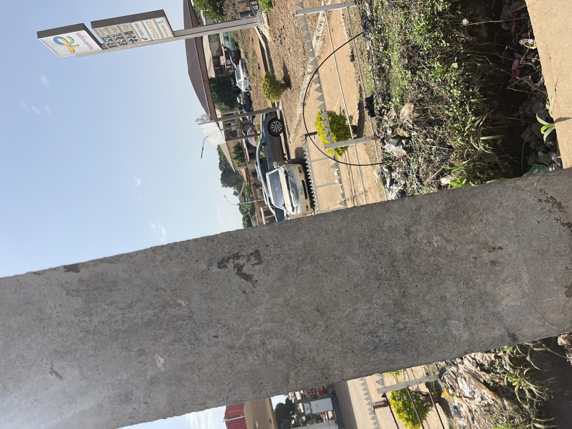 This image depicts a gas station in Bukuru, Nigeria, viewed from behind a large concrete pillar. The gas station appears to be branded as TotalEnergies, indicated by signage displaying the company logo and fuel prices. The prices listed are 134.9 for Petrol, 137.9 for Diesel, and 220.0 for Kerosine. Adjacent to the price board is signage for "TOUCH POINT" and "QUARTZ". Several cars are visible at the gas station, including a light-colored sedan parked in the foreground and a darker sedan. In the background, there are structures that appear to be part of the gas station or nearby buildings, along with trees and vegetation. The scene also shows a significant amount of litter and debris on the ground, particularly in the lower right portion of the image and along a ditch. The sky is clear and blue, suggesting a sunny day. The concrete pillar in the foreground is rough-textured and shows some signs of weathering.