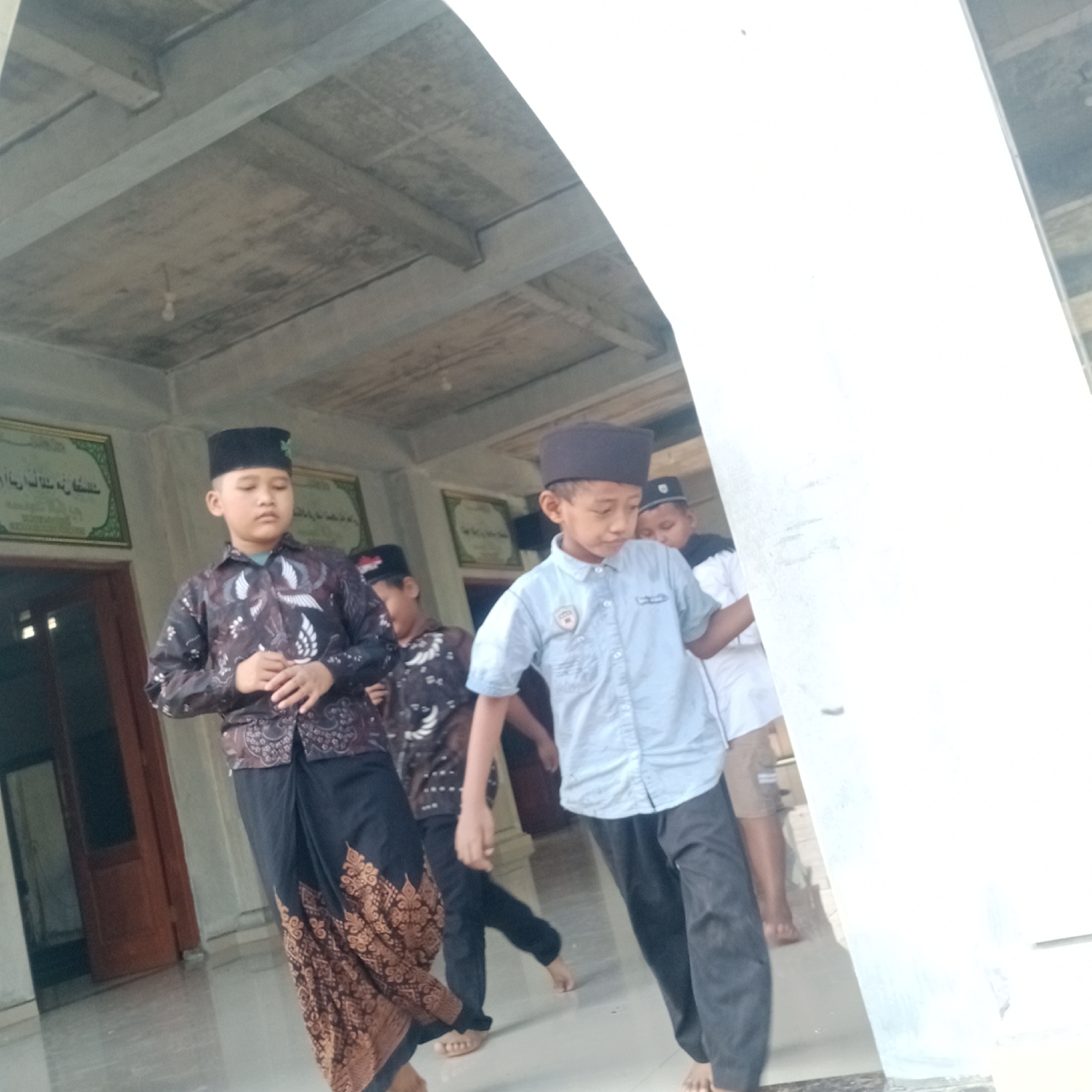 In Tegowanu Kulon, Indonesia, four young boys in traditional Islamic attire, including peci caps and sarongs, are seen walking within a building that appears to be a mosque or religious school. The boy in the foreground on the left wears a dark patterned shirt and a patterned sarong. To his right, another boy sports a light blue collared shirt and dark pants. Further back, two other boys, one in a dark patterned shirt and the other in a white shirt, are also visible. The environment features a concrete ceiling with visible beams, a light fixture, and what seems to be artwork or calligraphy displayed on the walls. The floor is tiled, and a wooden door is partially visible on the left side of the frame. The lighting suggests it is daytime.