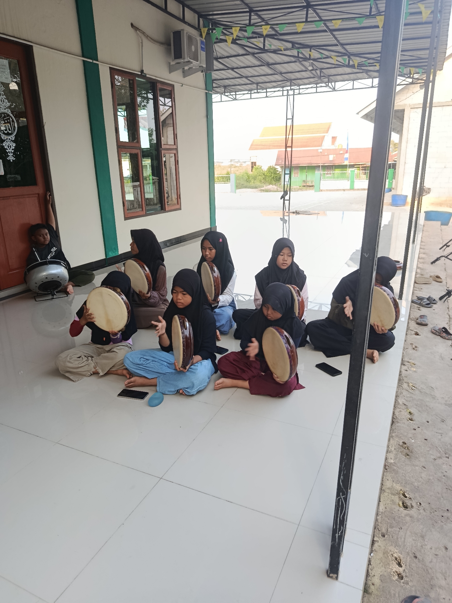 In Swarga Bara, Indonesia, a group of children is seated on a tiled floor outside a building, practicing music with tambourines. A young boy in a black t-shirt sits leaning against a doorframe, with a large circular drum-like instrument in front of him. Seven children, all wearing hijabs, are positioned in front of the building, each holding and playing a tambourine. The building has a white exterior with green trim around a wooden door and windows. A metal roof structure with triangular flags is visible overhead. The ground outside the tiled area is rough and textured. Several pairs of sandals and one open-toed shoe are scattered near the edge of the tiled area. A mobile phone and a blue oval object are on the floor among the children.