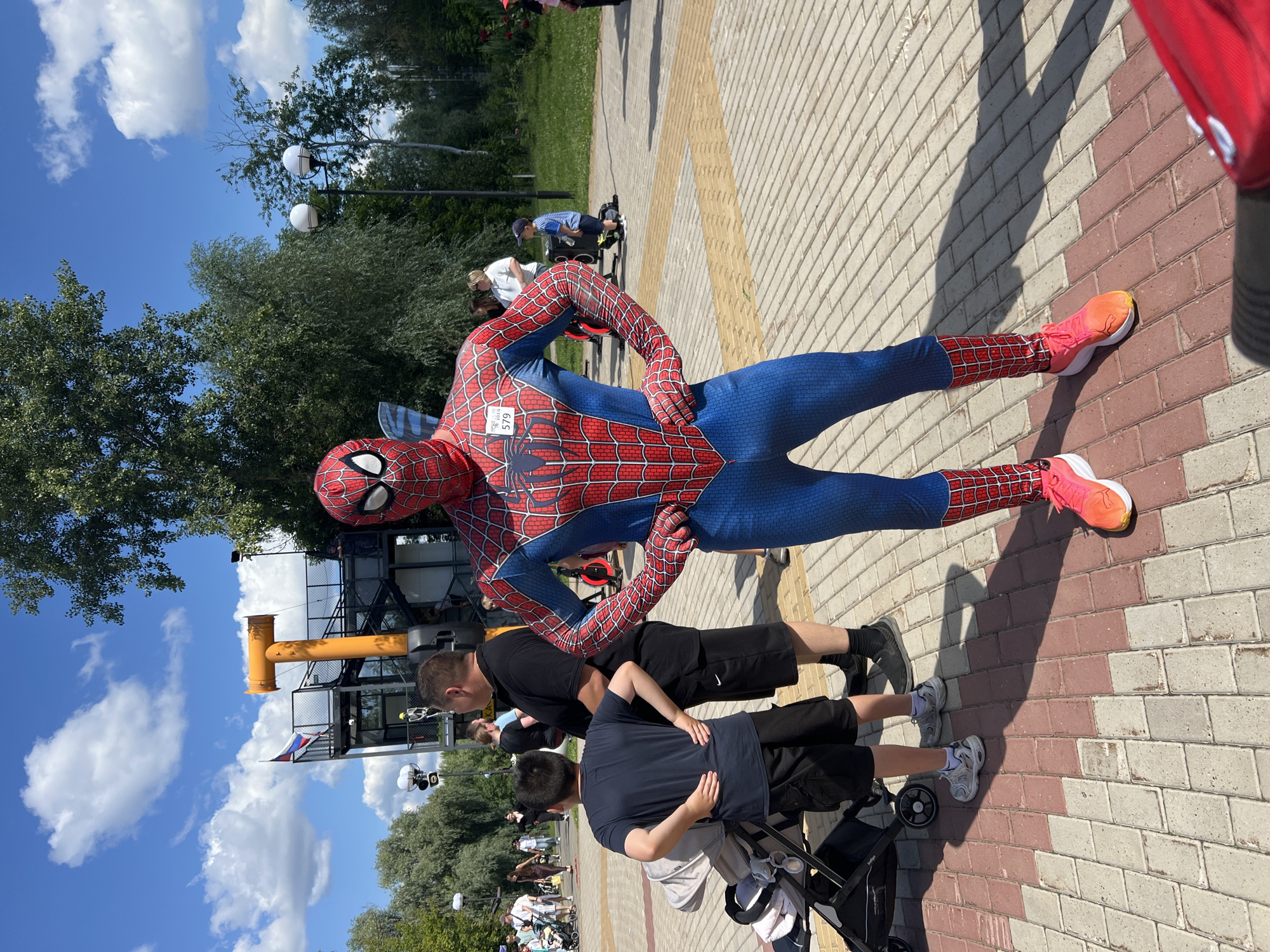 In an outdoor park setting, likely in Cheboksary, Russia, a person dressed as Spider-Man stands prominently in the foreground, facing the camera. The Spider-Man costume is a full-body suit in red and blue with the signature web pattern. On the chest of the costume, a small white tag with the number "579" is visible. The individual is wearing bright pink and orange athletic shoes.
To the left of Spider-Man, a young boy in a dark blue t-shirt and black shorts stands with his hands on his hips, looking away from the camera. He is positioned near a baby stroller. Behind the boy and slightly to his left, a man in a black polo shirt and black shorts is also visible, looking to the right.
In the background, other people are scattered throughout the park, some walking and others standing. Several trees and lampposts are present, suggesting a well-maintained public space. A distinctive yellow structure with a Russian flag flying on top is visible on the left side of the background. The ground is paved with reddish-brown and grey bricks arranged in a pattern. The sky is blue with scattered white clouds, indicating a sunny day.