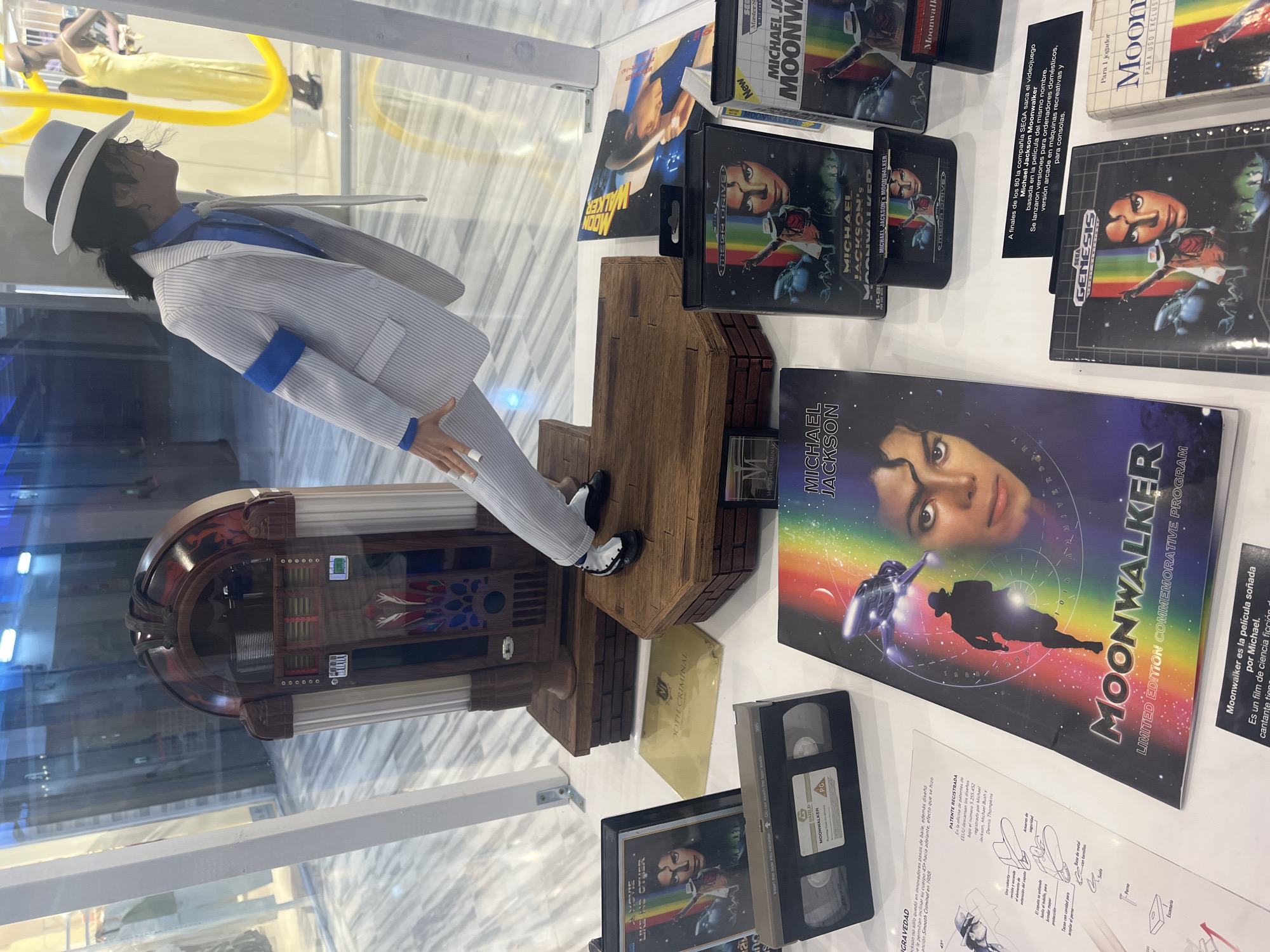 This image captures a Michael Jackson-themed exhibition display, likely housed within a museum or public venue in Las Palmas, Spain. The central focus is a life-sized or large figurine of Michael Jackson, dressed in a white pinstripe suit, blue shirt, blue armband, white fedora, and black-and-white shoes. The figure is posed in his iconic anti-gravity lean from the "Smooth Criminal" music video, supported by a wooden base that resembles steps, hinting at the patented shoes that allowed this illusion.
The display table, covered with a white surface, showcases an extensive collection of "Michael Jackson's Moonwalker" memorabilia. These include various video game cartridges for Sega Master System and Sega Genesis (Mega Drive), VHS tapes of the "Moonwalker" film (one with a "PG" rating), a large "Michael Jackson Moonwalker Limited Edition Commemorative Program," and other posters and media. One album cover for "Smooth Criminal" and a yellow item with "20th Criminal" are also visible.
Several information plaques in Spanish provide context. One explains that "Moonwalker es la película soñada por Michael," a science fiction film. Another describes how SEGA released the "Michael Jackson Moonwalker" video game in the late 80s for home computers, arcade machines, and consoles. A detailed plaque titled "PATENTE REGISTRADA" (REGISTERED PATENT) illustrates the mechanism of Jackson's anti-gravity lean shoes, highlighting the heel cavity designed to couple with a bolt, detailing the patent number 5.255.452, registered by Michael Jackson, Michael Bush, and Dennis Thompkins.
To the left of the figurine, a vintage-style jukebox, reddish-brown with silver accents, is partially visible within the display. The background shows reflections of other exhibits or people, indicating an active indoor environment.