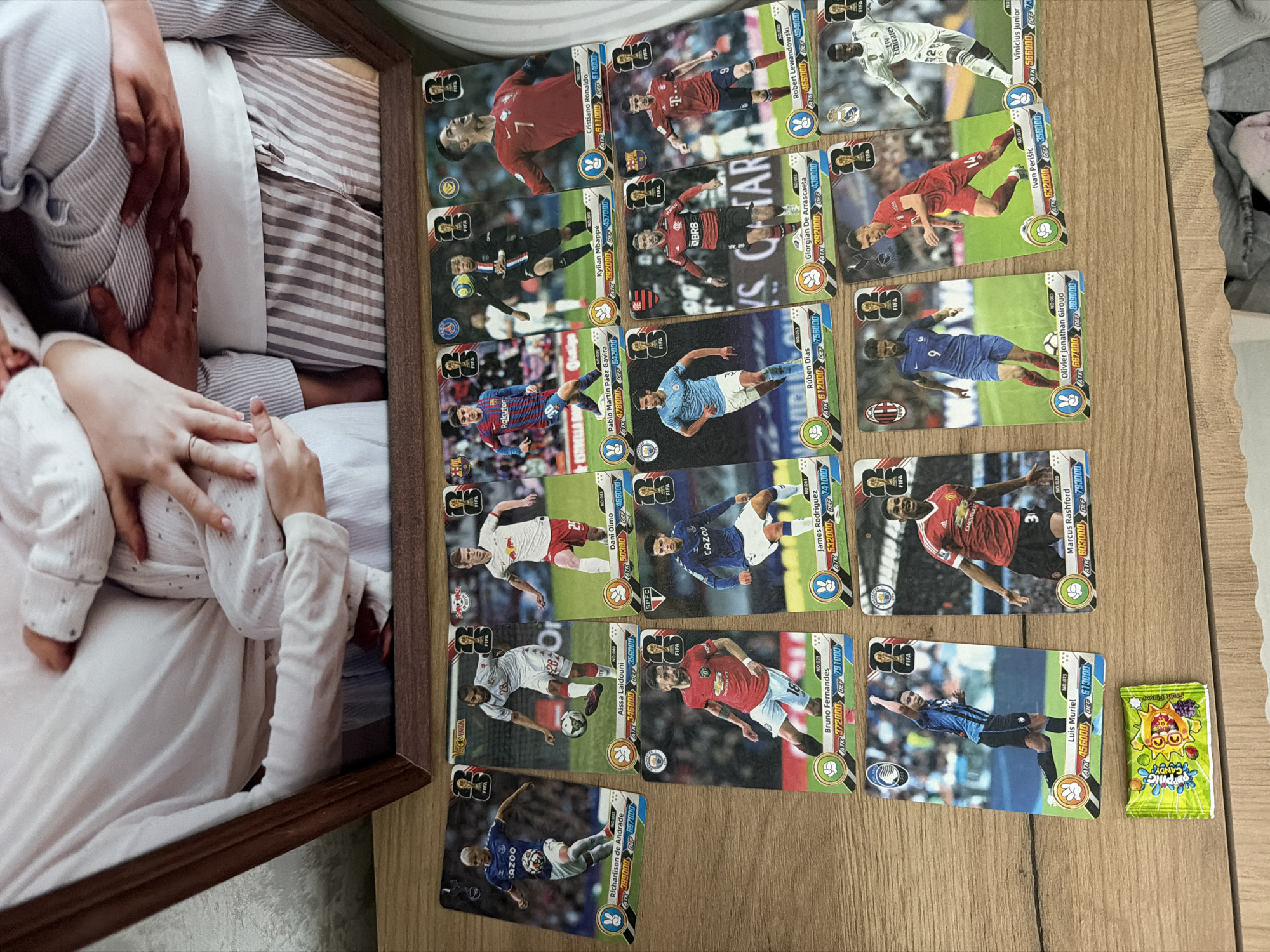 A framed photograph of an adult couple holding a baby is displayed at the top of the image. Below the frame, a collection of football trading cards is arranged on a wooden surface. The cards depict various football players in action, each with their name, number, and statistics printed on them. A small, green, foil-wrapped candy is placed to the left of the bottom row of cards. The scene is lit from above, casting shadows on the wooden surface. There is no visual information indicating a specific location in Kazan, Russia.