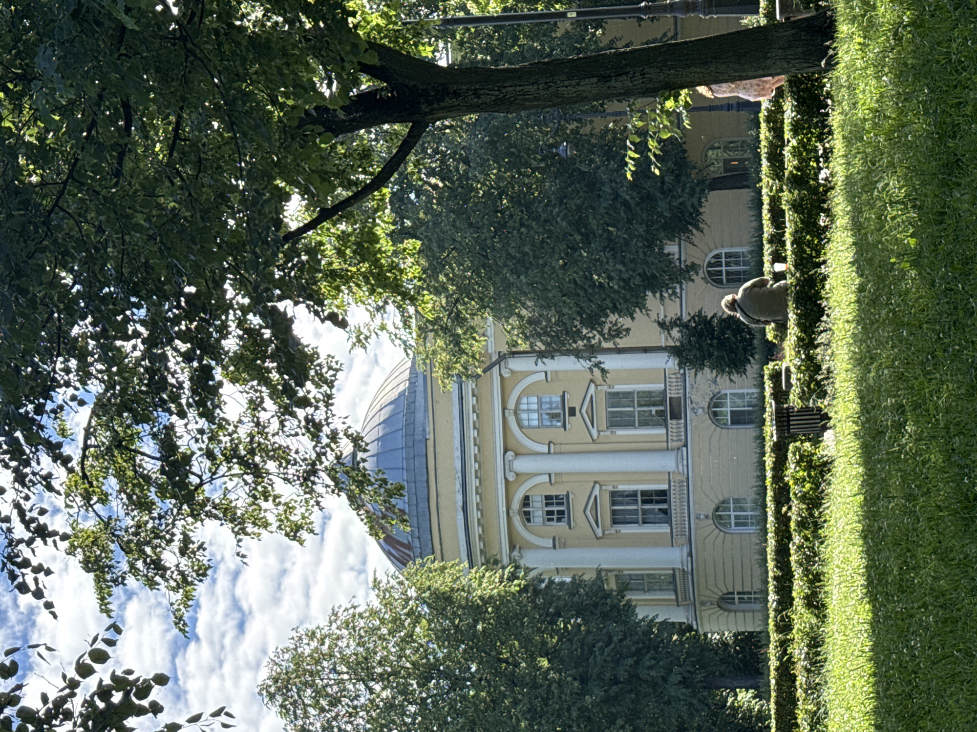 The image captures a sunny day in a park adjacent to a classical building with a dome in Saint Petersburg, Russia. In the foreground, lush green grass is dappled with sunlight. Behind the grass is a neatly trimmed hedge, and beyond that, a bench occupied by a person wearing a greenish-brown jacket, facing away from the camera. A plastic cup with a straw sits on the bench next to them. To the right of this individual, another person is visible, partially obscured by a tree trunk, wearing a pink floral dress and facing towards the building.
The building itself is a light yellow structure with a prominent dome. It features classical architectural elements such as large white columns and arched windows. Tall, leafy trees frame the building, casting shadows and creating a dappled effect on the scene. The sky above is blue with fluffy white clouds. The overall impression is a peaceful, summery scene in an urban park setting.