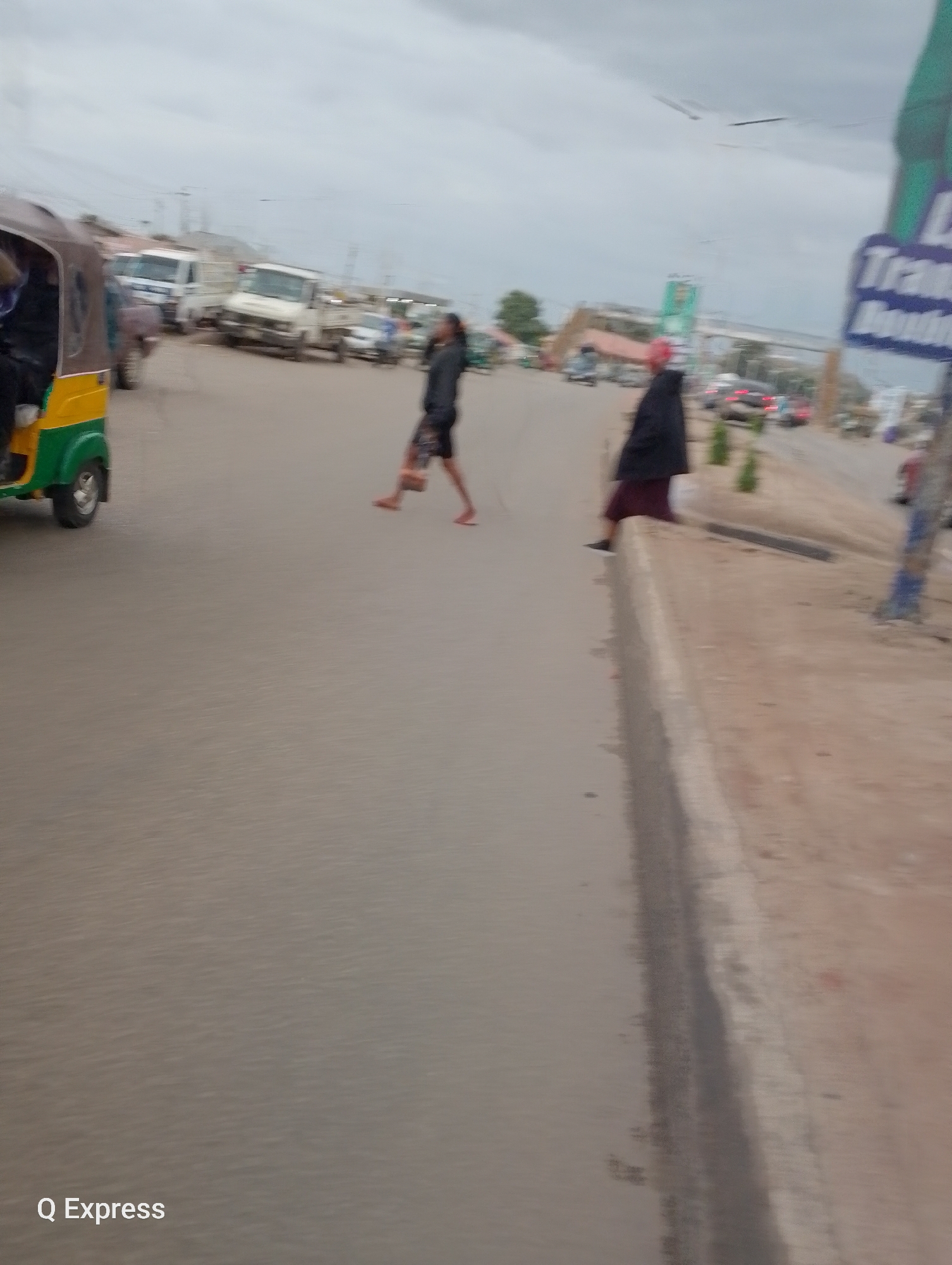 The image shows a street scene in Jos, Nigeria, with a focus on a pedestrian crossing. Two individuals are visible crossing the road. One person, wearing dark clothing and a red head covering, is walking from right to left. Another person, in dark shorts and a dark top, is walking in the opposite direction, from left to right, with their arms outstretched. A yellow and green auto-rickshaw is parked on the left side of the image. In the background, several vehicles, including white trucks and cars, are present on the road, suggesting moderate traffic. The sky is overcast, indicating cloudy weather. A sign with blue and white text, partially visible and blurry, is on the right side of the road. The road appears to be paved, and there is a concrete curb on the right side of the image. The overall scene suggests an ordinary day in a busy urban or semi-urban environment in Nigeria. The text "Q Express" is visible in the bottom left corner of the image.