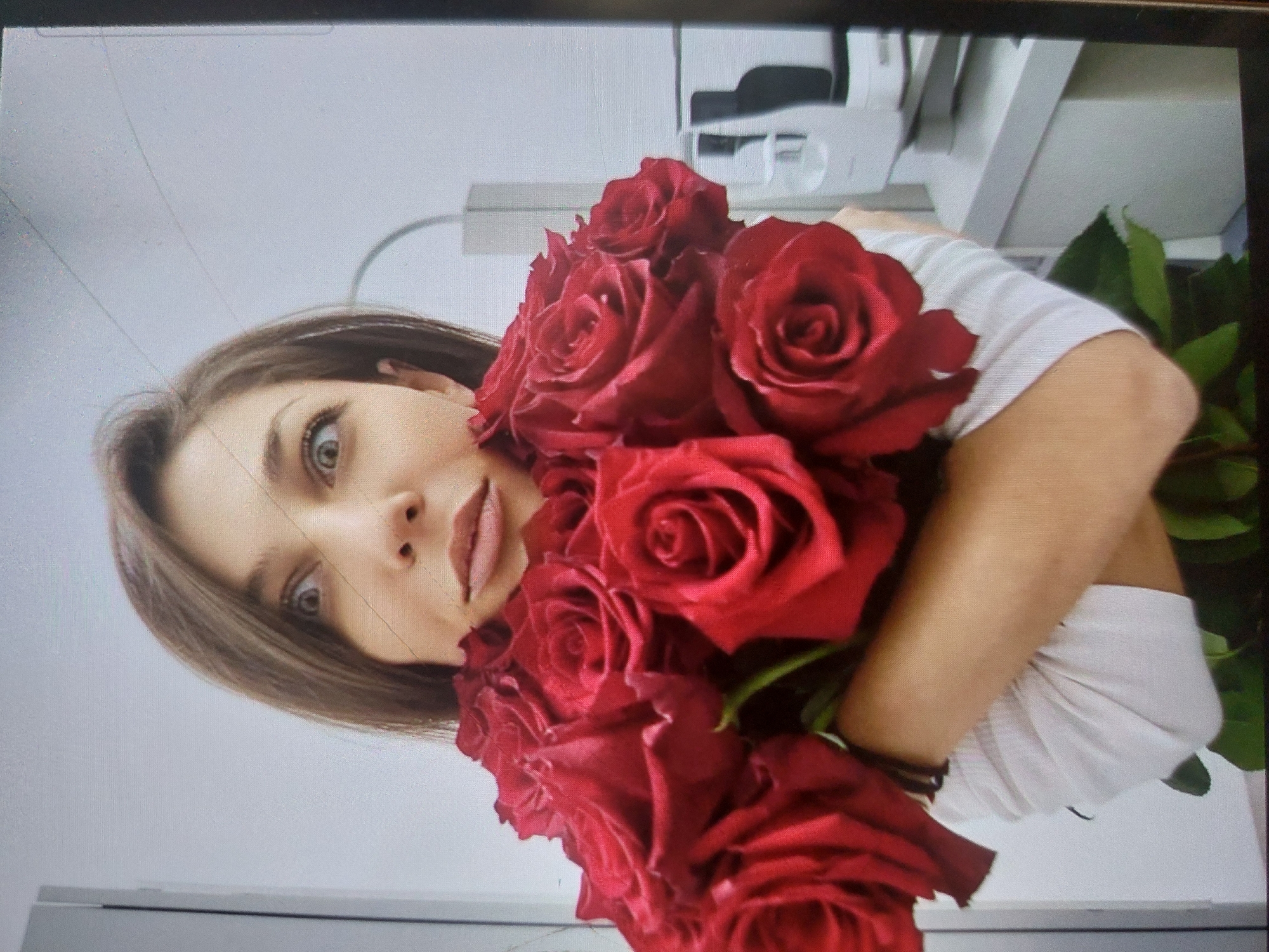 A woman is holding a bouquet of red roses. She has shoulder-length brown hair and is wearing a white t-shirt. The location appears to be indoors, possibly a doctor's office due to the medical equipment in the background. There's no verifiable indication that the image was taken in Tsarevo, Bulgaria, or that anyone is taking action or interacting with others. The woman is looking towards the camera. The environment is a mostly white room with various white fixtures.