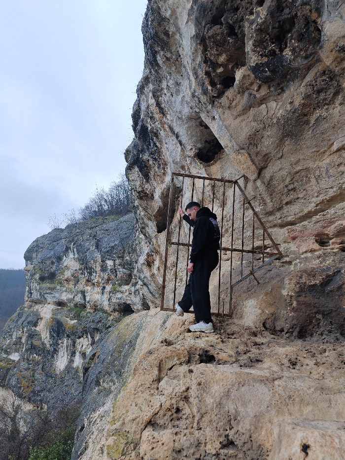 The image depicts a male individual standing on a narrow rock ledge within the Shumen Plateau, Shumen, Bulgaria, against a large, weathered rock face on an overcast day.
**Scene Description:**
The primary subject is a male, estimated to be an adult, positioned on an uneven, light-colored rock ledge. He wears a dark, long-sleeved hooded top with a white graphic on the upper back, dark trousers, and white athletic shoes. His hair is short and dark. He is oriented towards the left side of the frame, with his head tilted downward and his left arm bent, resting his forearm on the top horizontal bar of a rusted, brown metal gate structure. This gate, composed of vertical and horizontal bars, appears crudely constructed and is partially affixed to the main rock face directly behind the subject. The gate's design includes a taller vertical section and a shorter section angled upward and to the right.
The rock face itself, occupying the right side and extending into the background, is a prominent geological feature, exhibiting varying textures, erosion patterns, and colorations ranging from light beige to dark grey and black. Multiple natural openings, indentations, or small cave entrances are visible within the rock, one directly behind the gate structure.
In the left background, another large, rugged cliff face is visible, sparsely covered with leafless deciduous trees and bushes. The sky above is uniformly overcast and light grey. The overall environment is rugged and natural, suggesting a high-elevation or mountainous terrain.