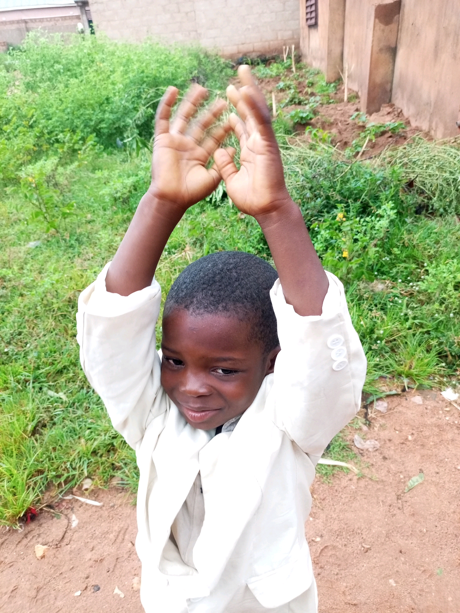 A young boy with dark skin and black hair is captured in a medium shot, standing outdoors. He is wearing a light-colored suit jacket with three buttons on the cuff. His arms are raised above his head, and his hands are clasped together, palms facing the camera. The boy is looking towards the camera with a slight smile.
The background consists of lush green foliage, a dirt ground, and a portion of a light brown building. The building appears to be constructed with brick and has a simple, utilitarian design. Some small plants are visible in a cultivated patch near the building. The overall lighting suggests it is daytime. The image appears to have been taken in a rural or semi-urban area in Tempure, Nigeria.