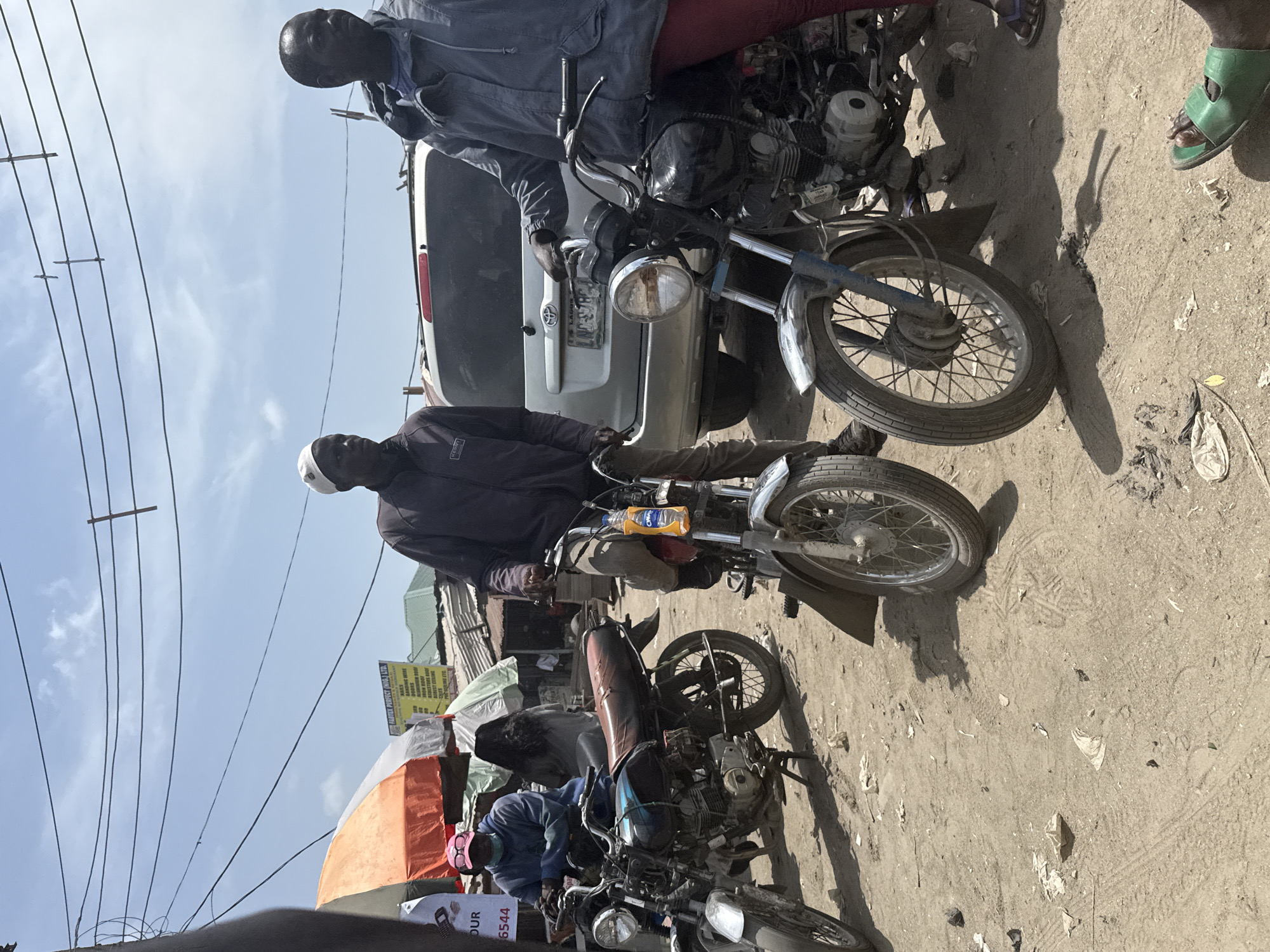 The image depicts a bustling, unpaved street scene in a commercial area of Lagos, Nigeria, identifiable by the "LND" license plate on a parked SUV and the "FASTER PERFECT (NIG.) LTD." business sign. The environment is dusty, under a bright, partly cloudy sky, with numerous overhead electrical wires.
In the foreground, three dark-skinned males are present. The central figure, wearing a dark jacket and a light skullcap, stands between two motorcycles. He holds the handlebars of a motorcycle to his right, from which a yellow "O'WAP" bottle hangs. To the far right, a second male, wearing a grey/blue hooded jacket and reddish trousers, holds the handlebars of a foreground motorcycle; his right foot in a green slide sandal is visible. In the middle ground, a third individual, clad in a blue hoodie and pink cap, is seated on a third motorcycle. Behind them, partially obscured, is a person with long dark hair.
Several objects populate the scene: three visible motorcycles, all appearing to be older models, one with a worn reddish-brown seat. A silver Toyota SUV (license plate LND-378CH) is parked in the background. Overhead, a dense network of black electrical wires crisscrosses the sky. The ground is unpaved, consisting of dirt and sand, marked by tire tracks and scattered debris including plastic and paper. Informal structures with corrugated metal roofs and two market umbrellas (one orange and white, one light green) are visible in the background, along with a yellow business sign for "FASTER PERFECT (NIG.) LTD." listing various construction materials. The bright daylight casts distinct shadows.