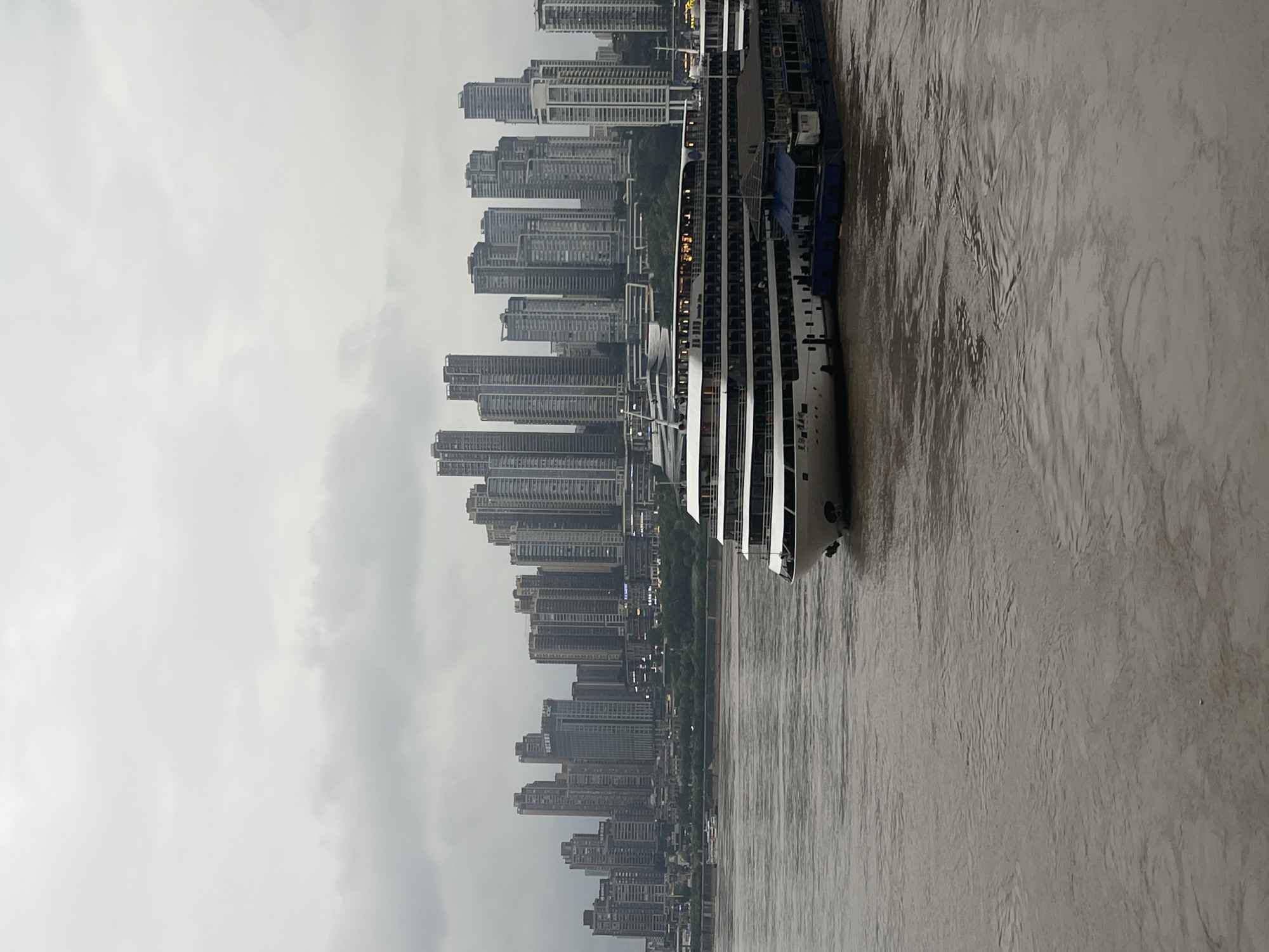 The image captures a cloudy day in the Yuzhong District of Chongqing, China. A large white cruise ship is docked in the foreground, with several decks visible and illuminated windows suggesting activity on board. Moored alongside the cruise ship are smaller dark blue barges. The murky brown waters of a river, likely the Yangtze, flow in front of the vessel. In the background, a dense cityscape of tall, modern buildings rises from the riverbank, showcasing a mix of architectural styles and heights. The sky is overcast, contributing to a subdued, diffused light across the entire scene.