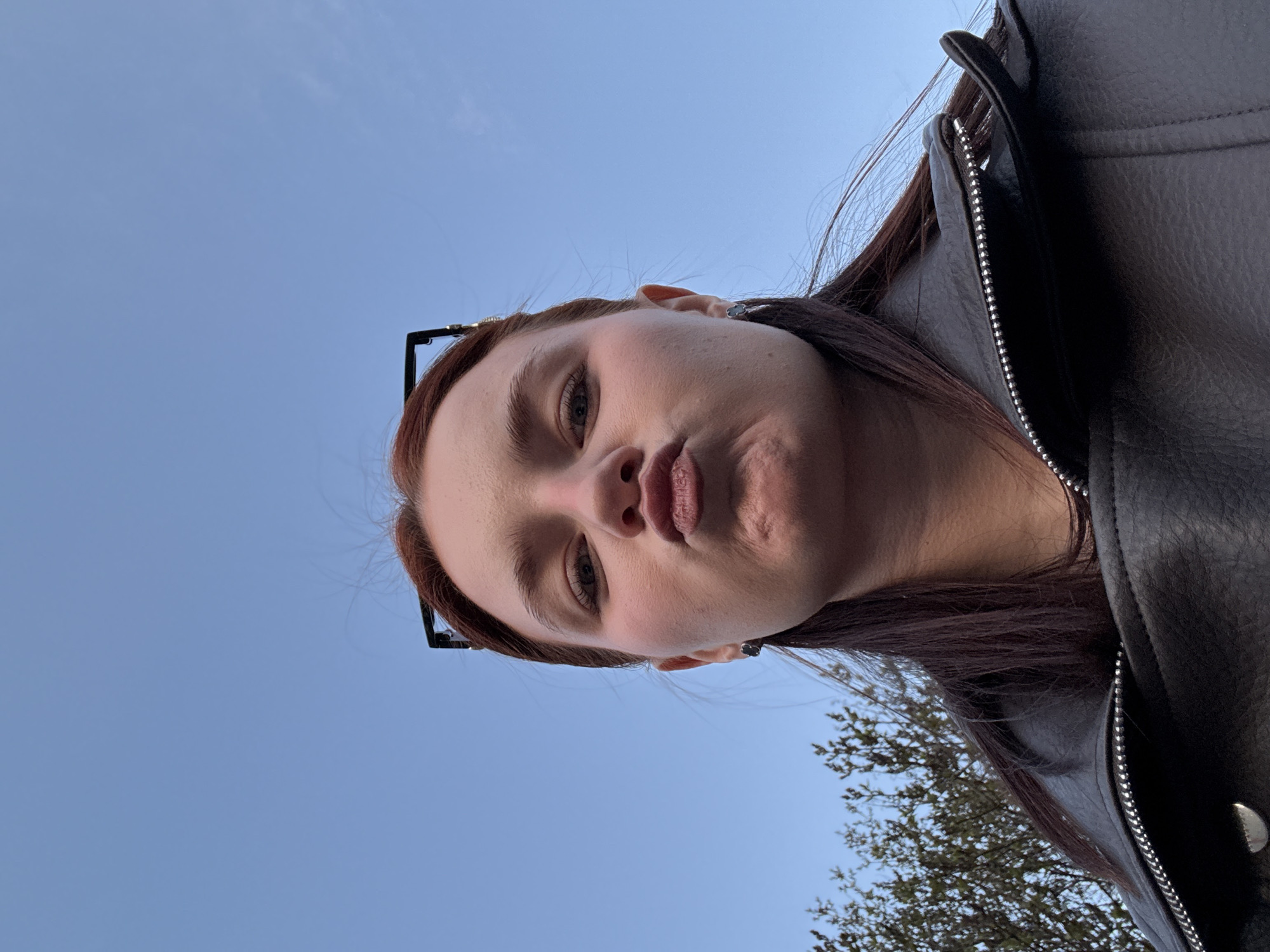 A selfie of a person with light skin, auburn hair, and pursed lips. The person wears a black leather jacket and glasses. The background consists of a blue sky and a partial view of a tree with bare branches. The image was taken from a low angle, looking upward. The location is not discernible as Saint Petersburg, Russia based on the visual information provided.