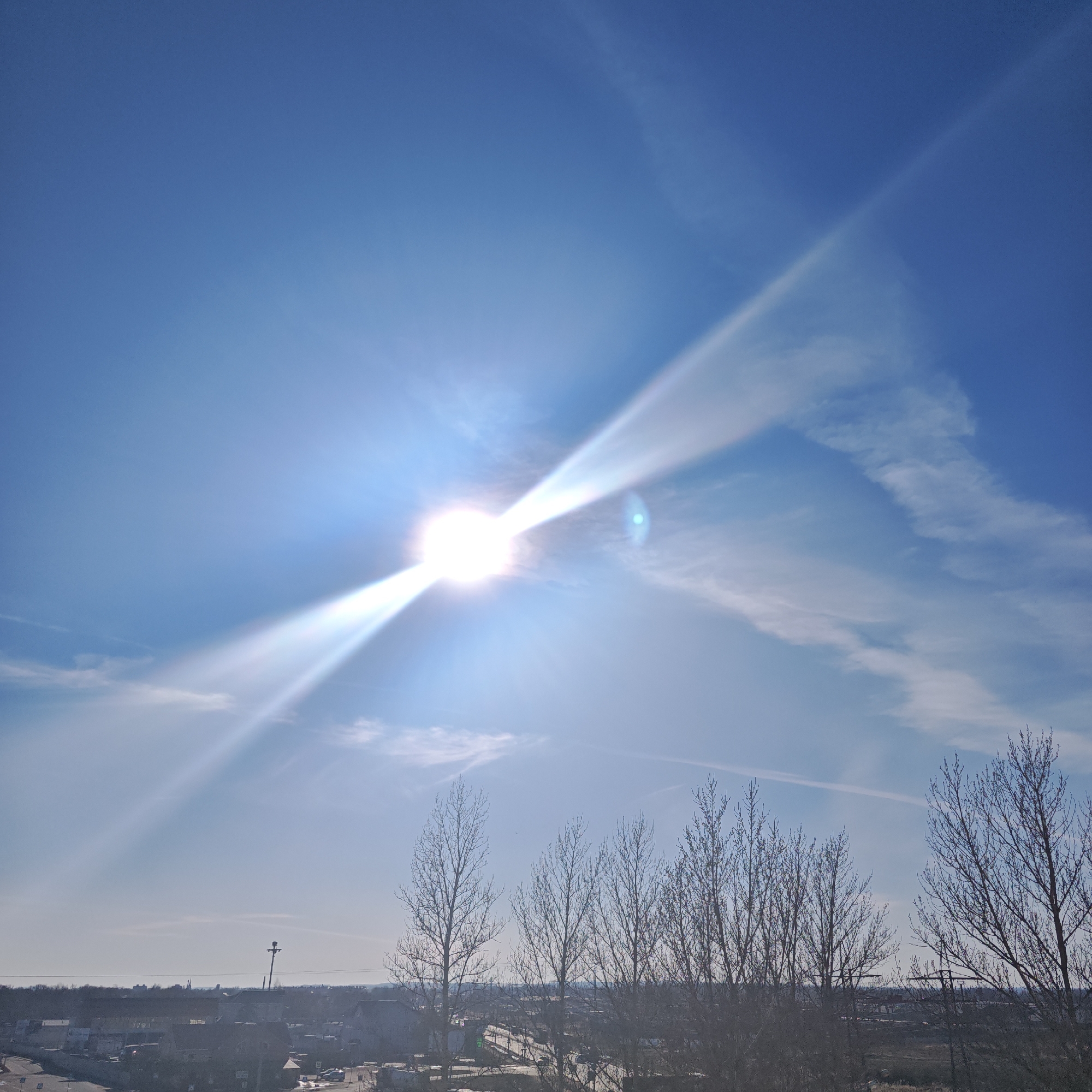 The image shows a bright, sunny day in Rostov, Russia. The sun is high in the sky, radiating strong beams of light. Bare trees are silhouetted in the foreground. Below them is an urban landscape with buildings and roads. A utility pole can be seen in the distance. The sky is a clear blue with some wispy clouds.