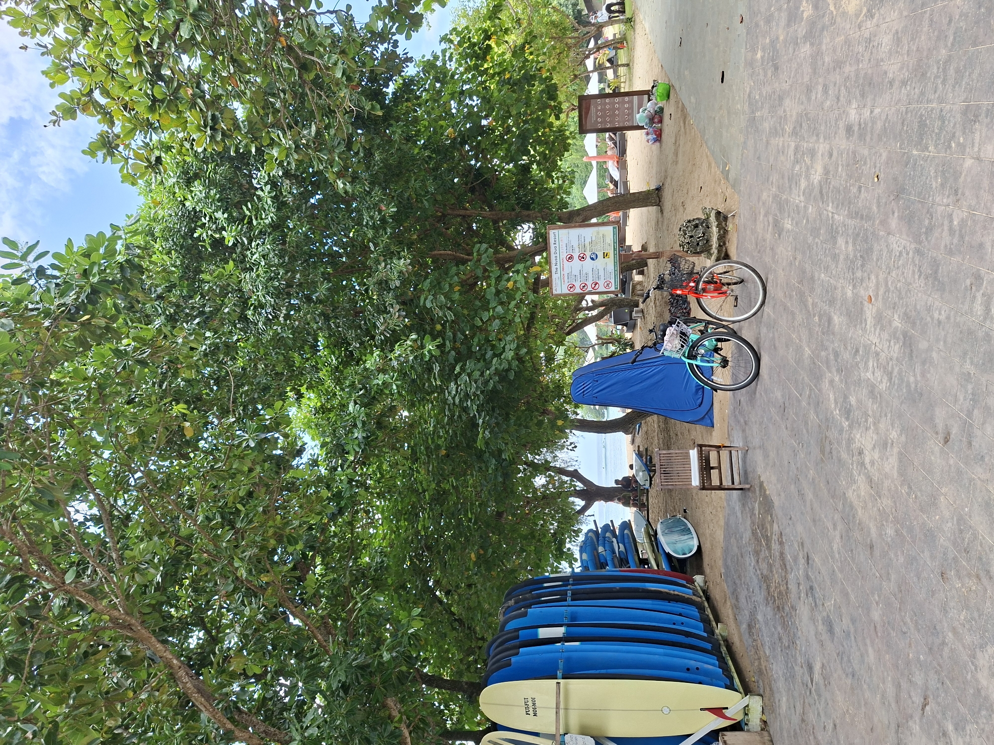The image presents a scene along a concrete pathway in Kuta Selatan, Indonesia, near the beach. The pathway is lined with a dense green tree on the left, partially obscuring the sky.
Along the edge of the path, adjacent to the sandy beach area, there is a stack of surfboards of various colors, primarily blue, with a beige one at the front. Adjacent to the stack, is a wooden chair. Further along the path is a blue beach tent and two bicycles are parked near signs with information about the beach. The bicycles are red and turquoise. In the background, there are additional people visible on the beach under umbrellas. The beach itself leads to the ocean in the distance.
