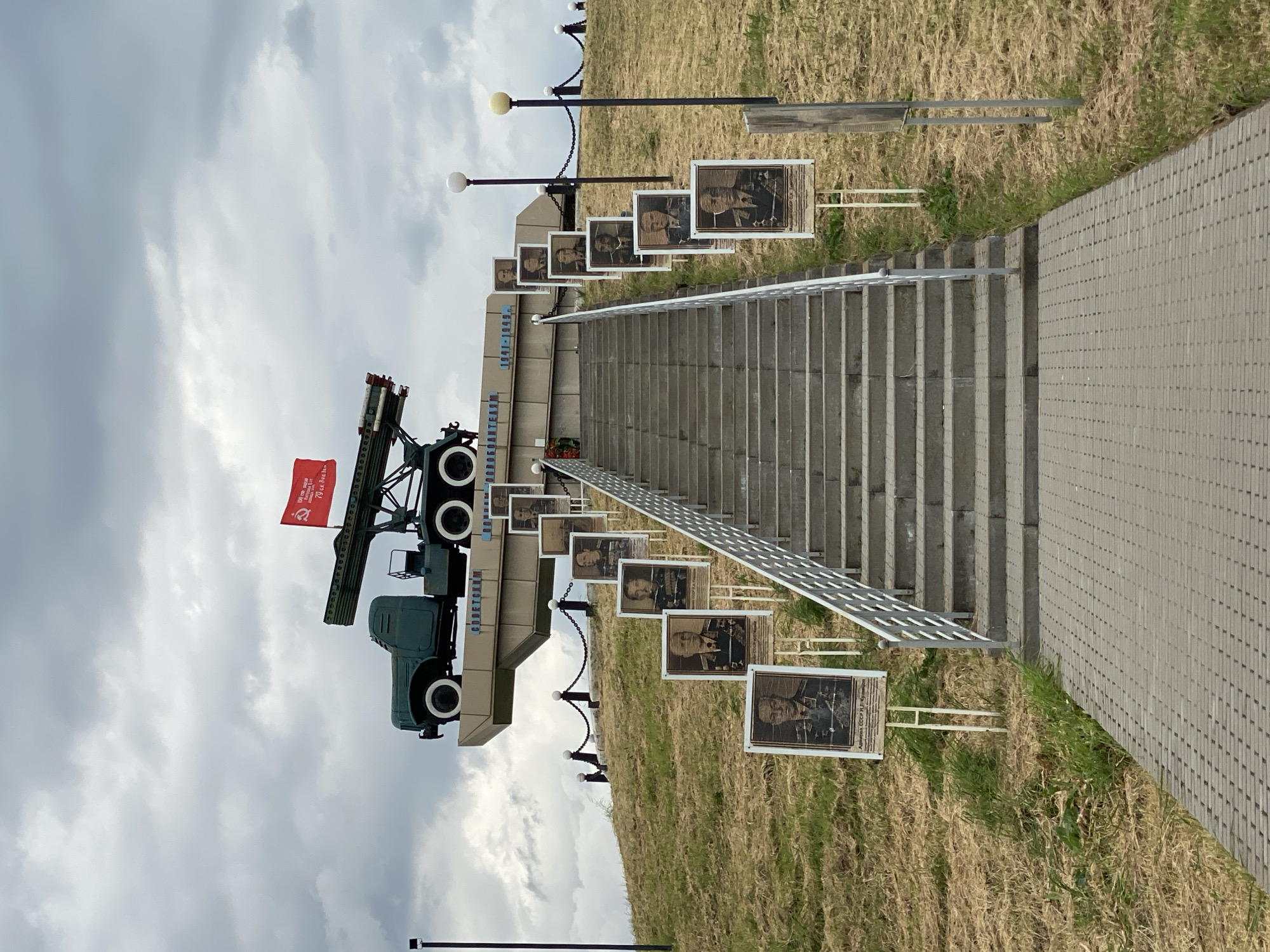 The image depicts a staircase leading up a grassy hill. Alongside the staircase are framed portraits on stands. At the top of the staircase is a Soviet-era missile launcher on a truck. The location appears to be a monument or memorial. The sky is overcast. Based on details visible on the monument, this may be in Bryukhovetskaya, Russia.
