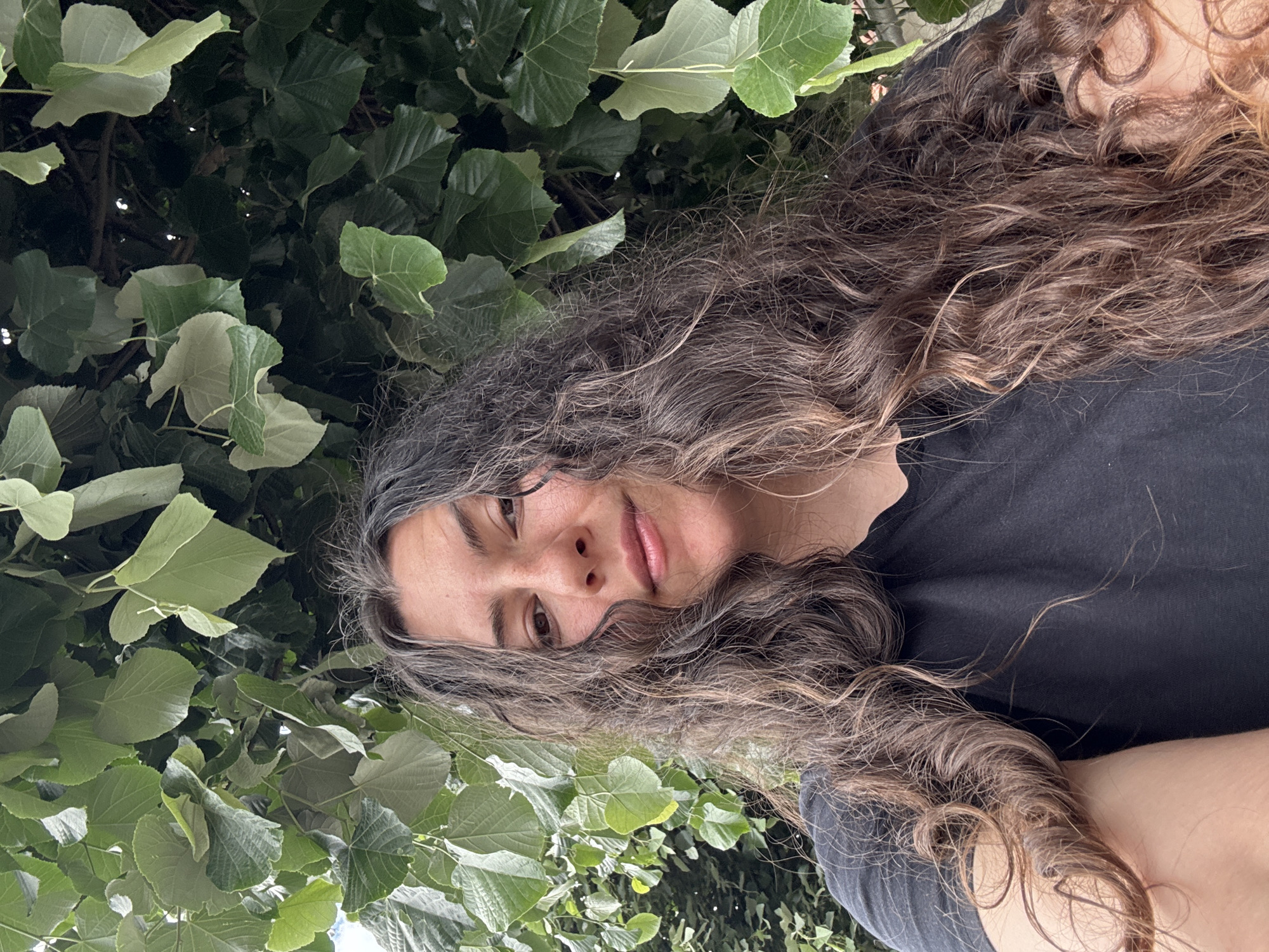 A person with long, curly, brown hair is centered in the image, wearing a black t-shirt. Green leaves surround the person, suggesting an outdoor location. The specific location within the city of Unknown, Unknown is not identifiable from the visual information.