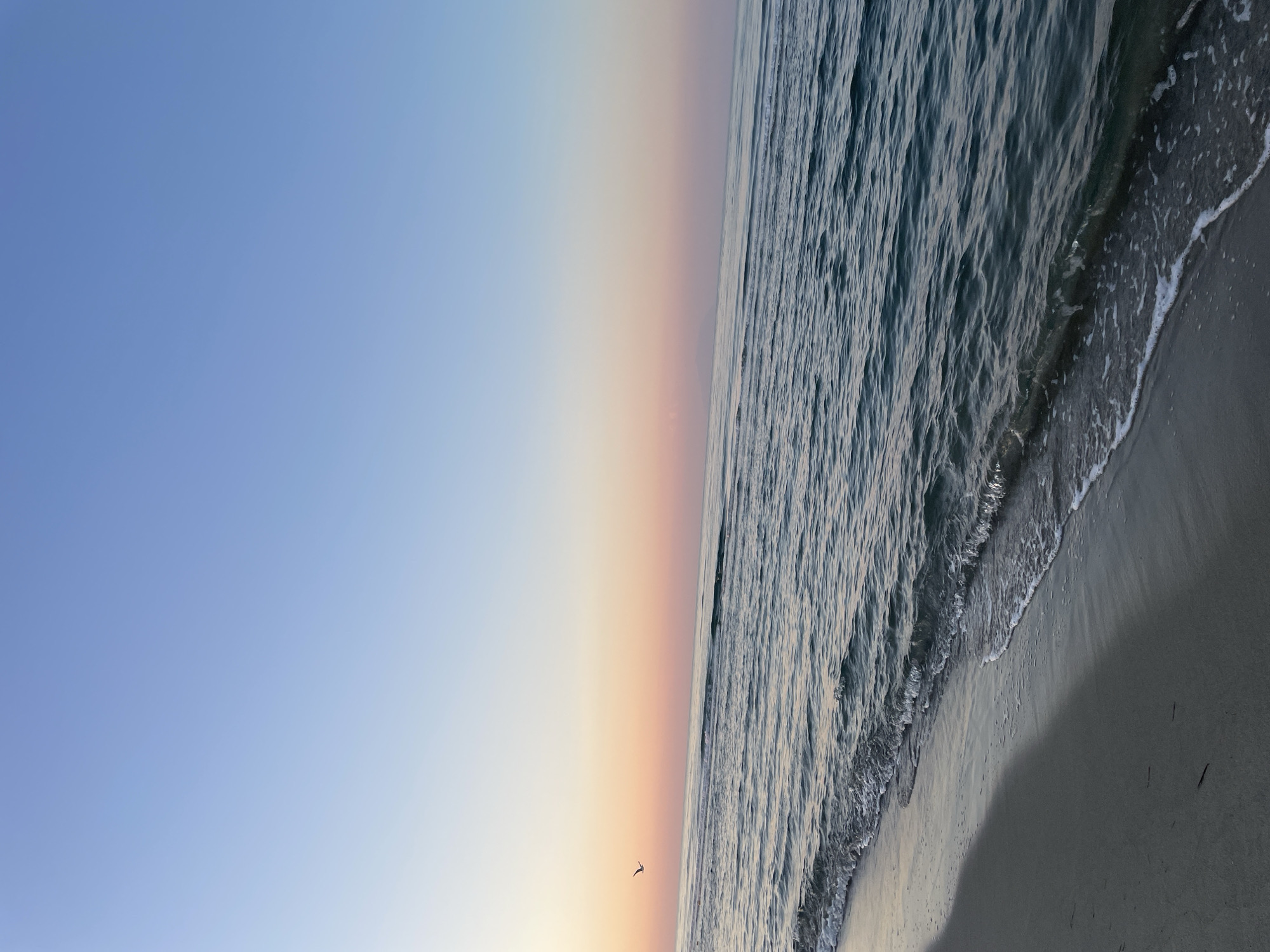 The image depicts a serene seascape at sunrise or sunset. The sky transitions from a pale blue at the top to soft pink and orange hues near the horizon, creating a gradient effect. A single bird is silhouetted in flight against the colorful sky. The ocean occupies the majority of the frame, with gentle waves rolling towards a sandy shore. The water is a deep blue-green, reflecting the colors of the sky. The foreground shows a sandy beach, wet from the receding tide, with subtle shadows cast by unseen elements. The overall scene evokes a sense of tranquility and natural beauty. The location is described as Unknown, Greece.