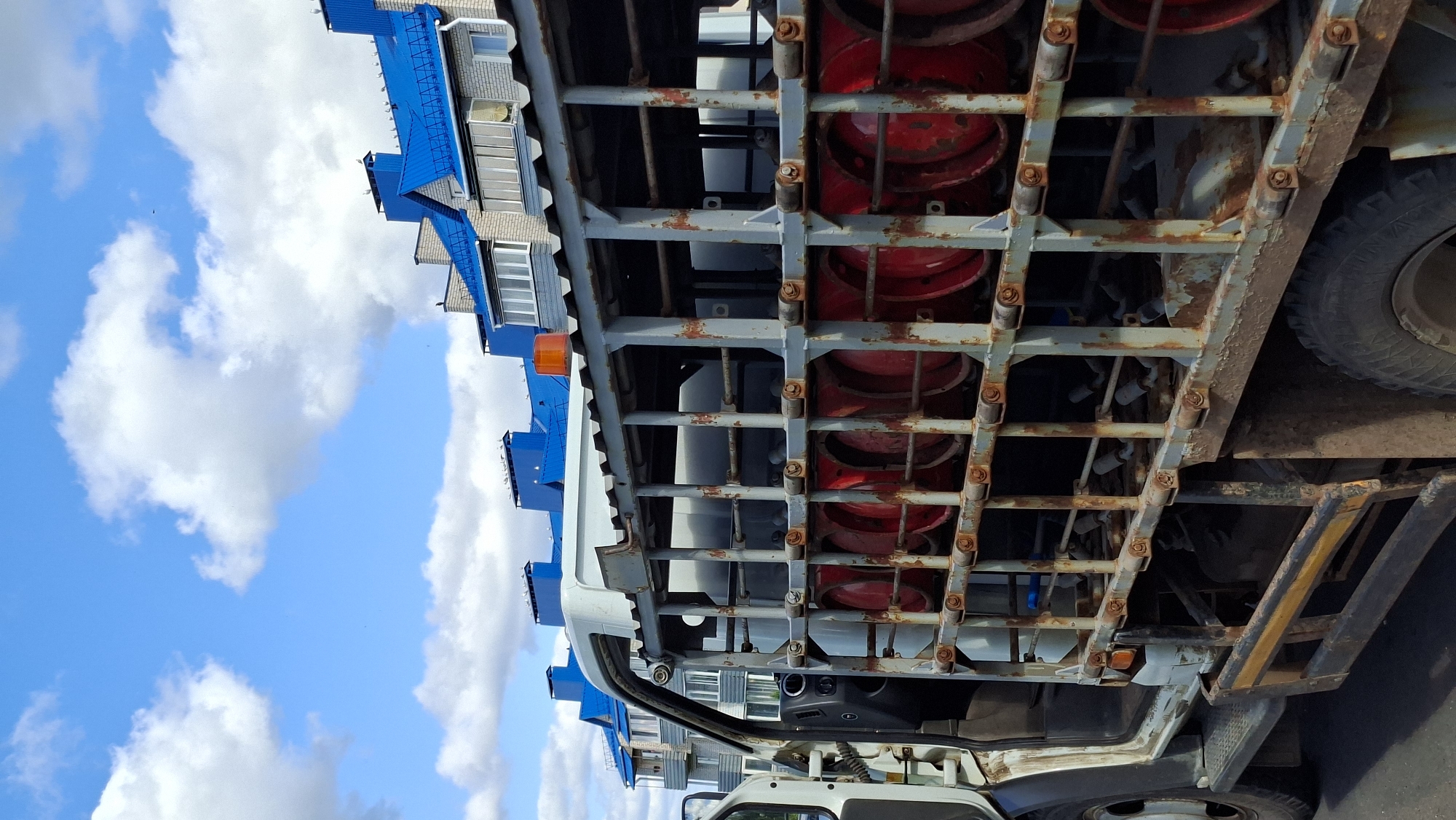 A vertically oriented, full-shot, eye-level, medium-distance photograph captures a weathered, light-colored truck loaded with red propane tanks. The truck, positioned on an asphalt surface, is a commercial vehicle with an open driver's side door revealing a black dashboard. The rear of the truck features a grey, metal cage structure holding the propane tanks, which are arranged in two rows, partially visible. An orange flashing light is mounted on the top of the cage. In the background, a modern building with a distinctive blue roof and grey brick facade stands against a bright blue sky dotted with white, fluffy clouds. The scene appears to be a street or roadside location in the city of Myadzyel, Belarus.