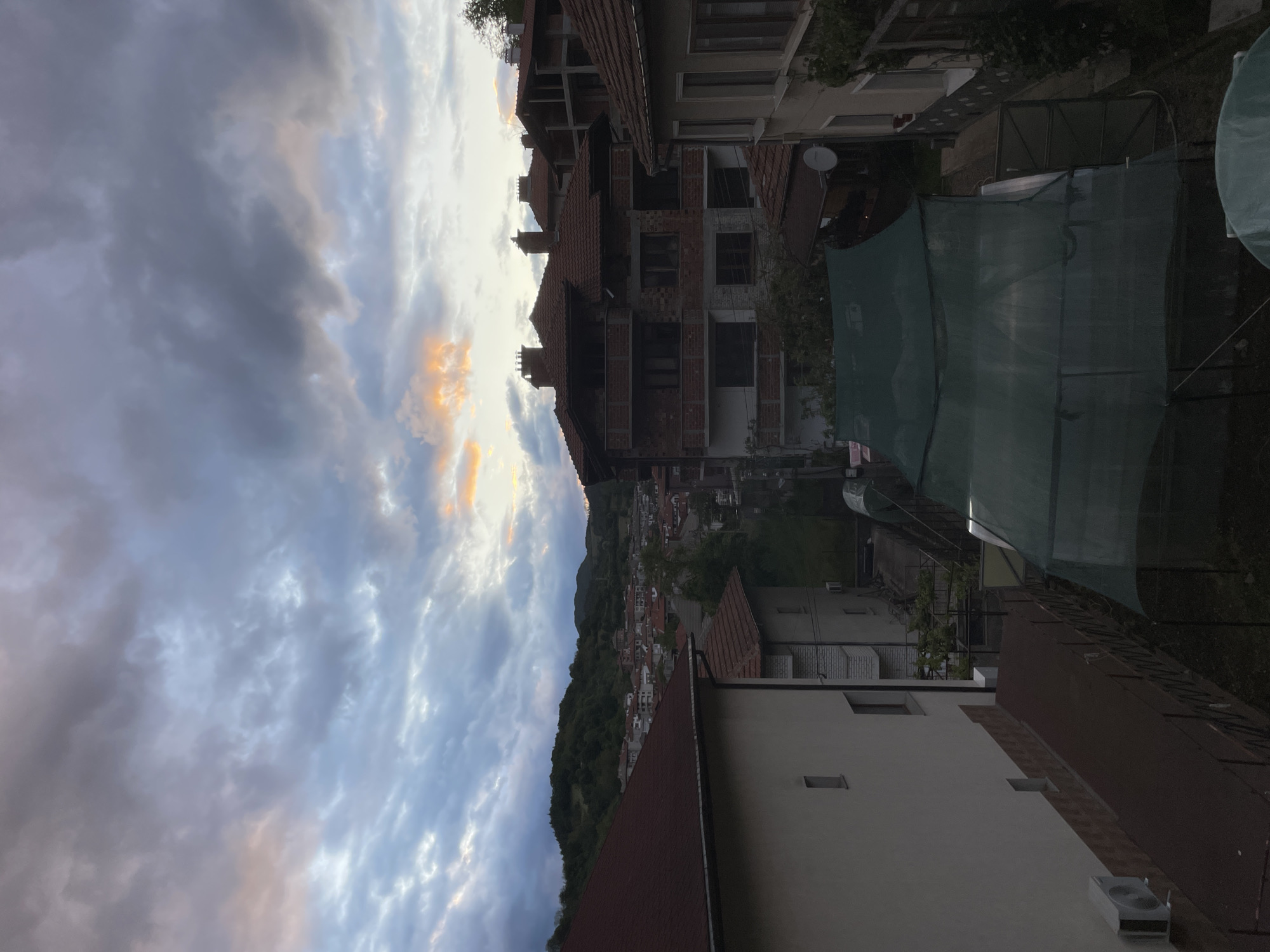 The image shows a view in Zlatograd, Bulgaria, at dusk or dawn. The foreground is dominated by buildings with red-tiled roofs and light-colored walls. In the middle ground, there are buildings with multiple stories, brick construction, and partially constructed sections. Beyond the buildings, there's a view of the town nestled in a valley, surrounded by tree-covered hills. The sky is filled with clouds tinged with hues of orange and blue. No people are visible. The lighting suggests either sunrise or sunset. A green mesh-like barrier or covering is visible in the foreground to the right. A satellite dish is mounted on one of the buildings.