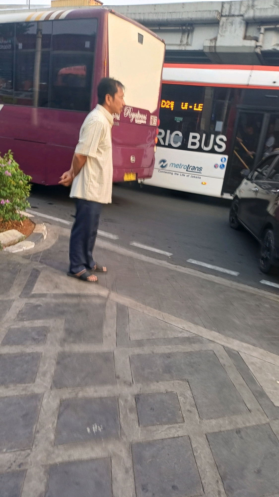 A man stands on a sidewalk, facing left, with his hands clasped behind his back. He wears a light-colored, short-sleeved collared shirt and dark pants. He is barefoot in sandals. Behind him, a maroon bus is parked, and to its right, a white Metrotrans bus displays "D21 U-LE" and "RIC BUS" on its side. The Metrotrans bus also shows the text "Connecting the Life of Jakarta." A black car is visible to the right of the white bus. The ground is paved with gray square tiles. A small bush with pink flowers is to the left of the man. The scene appears to be a bus stop or transit area in South Jakarta, Indonesia, likely during daylight hours based on the lighting.