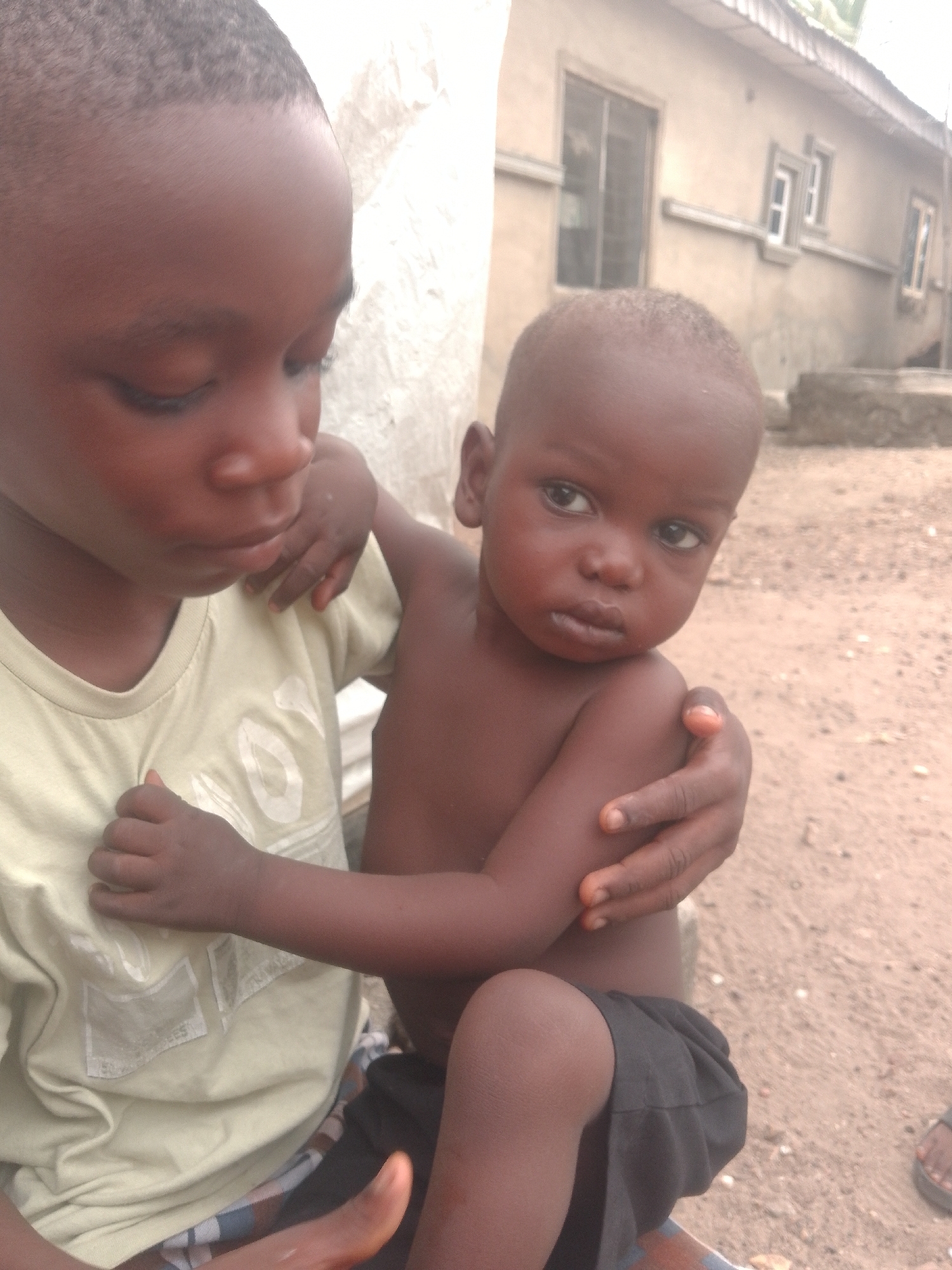 The scene depicts two children outdoors in the city of Unknown, Unknown. In the foreground, a dark-skinned child, identified as older, wears a light greenish-yellow short-sleeved t-shirt with faded white graphics and patterned shorts. His gaze is directed downwards, and his left arm is wrapped around the back of a younger, dark-skinned toddler. The toddler is nude from the waist up, wearing dark shorts, and is seated. His left arm is around the older child's waist, and his right hand rests on the older child's left arm. The toddler looks directly forward with a neutral, slightly pursed-lip expression. The background features a light-colored, single-story building with a dark corrugated metal roof. The building exhibits a large rectangular window with internal bars on the left and two smaller, framed windows on the right, with a horizontal decorative band running along the wall. The ground beneath the subjects is light brown dirt or sand mixed with small pebbles. A dark-colored sandal or flip-flop is partially visible on the far right of the frame.