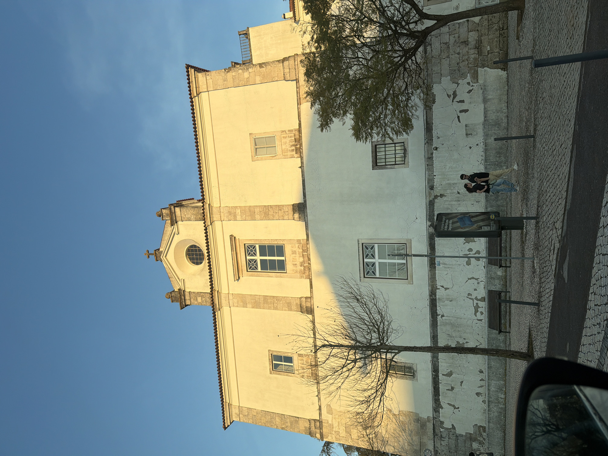 This image depicts the exterior of the Igreja do Seminário de Santarém, also known as the Church of Our Lady of Piety, located in Santarém, Portugal. The perspective is a low-angle shot from street level, likely from a vehicle, with a blurred side mirror visible in the bottom-left foreground.
The prominent structure is a large, multi-story historic building with an off-white/pale yellow stucco facade. The upper portion of the facade is brightly illuminated by direct sunlight, while the lower portion is partially in shadow. The building features classical architectural elements, including stone pilasters, a prominent cornice, and a central pediment topped with a stone cross. A circular oculus window is set within the pediment. Multiple rectangular windows of varying sizes are present across the facade; some have white frames and multiple panes, while others display dark grilles. Traditional terracotta barrel roof tiles are visible along the building's top edge. The lower half of the facade exhibits significant wear, with peeling stucco revealing underlying stone blocks and rough plasterwork.
The foreground consists of a cobblestone pavement with a narrow strip of asphalt in the immediate foreground. Several dark metal bollards delineate the edge of the paved area. Two dark brown wooden park benches are situated on the sidewalk. A black advertising billboard, partially displaying "IKEA" text, stands on the left side of the sidewalk.
Two individuals are walking from left to right on the cobblestone sidewalk in the mid-ground. The person on the right is male, wearing dark trousers, a dark long-sleeved shirt, and sunglasses. The person on the left is female, wearing light blue jeans and a dark short-sleeved top. They are walking side-by-side.
Two trees are present: a leafless deciduous tree with bare branches stands in the left-center, casting a shadow onto the building's lower wall. A second tree with green foliage is positioned on the right, partially obscuring a window on the building. The sky is clear and deep blue, occupying the upper half of the frame. The strong directional light and distinct shadows indicate either early morning or late afternoon.