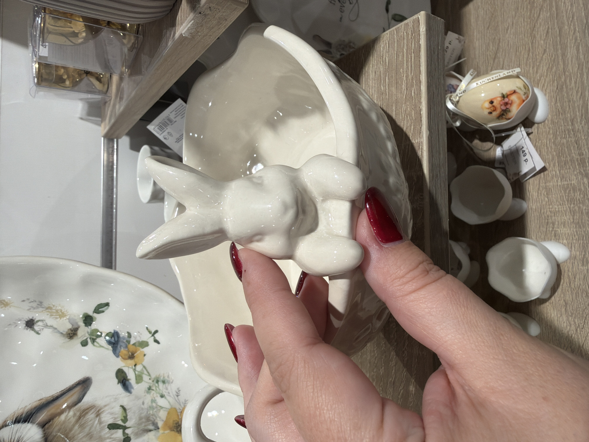 The image shows a person's hand with dark red nail polish holding a white ceramic dish with a bunny-shaped handle. The dish is part of a set, with a floral-patterned plate visible to the left. In the background, there are various decorative items, including small porcelain eggs and additional white ceramic pieces. The scene appears to be inside a retail store in Kazan, Russia, based on clues such as the tags with the price "149 P." (likely referring to Russian Rubles), and the item "KUCHENLAND" suggesting a brand or shop present in Russia. The person is likely inspecting or displaying the dish.