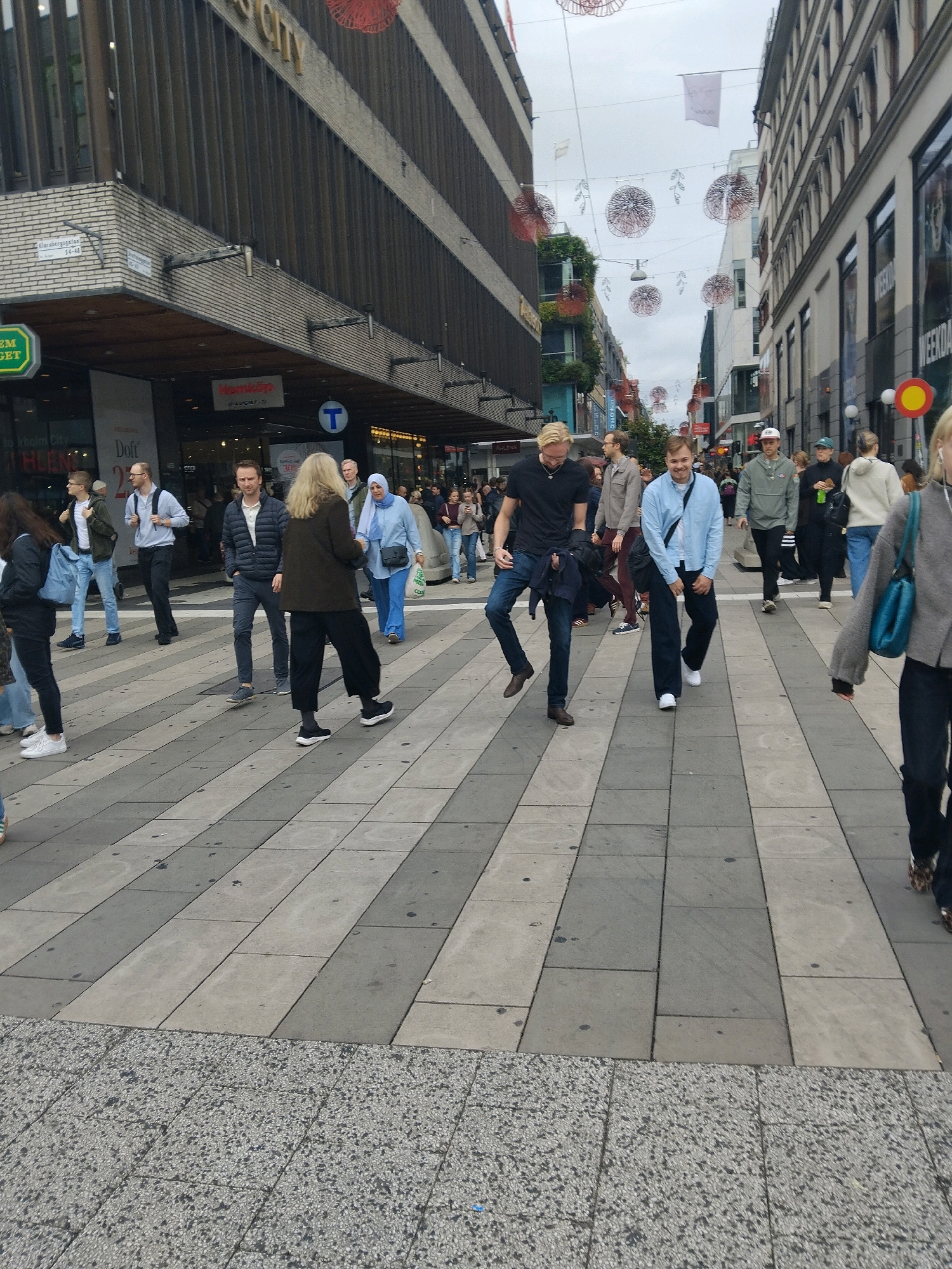 The image depicts a street scene in Stockholm, Sweden, likely during daytime. Several individuals are walking on a pedestrian walkway paved with alternating light and dark gray rectangular tiles. In the background, there are multi-story buildings with storefronts.
On the left side, a man with blonde hair wearing a dark t-shirt and jeans walks towards the viewer, carrying a dark jacket over his left arm. To his right, a man in a light blue collared shirt and dark pants is walking away from the viewer, with one leg bent as if mid-stride. He is smiling. Further back and to the left, a woman in a dark jacket and black pants walks away from the viewer, and a person in a light blue hijab and dark pants is walking across the path.
On the right side of the image, a woman wearing a gray jacket and dark pants is walking away from the viewer, her back mostly to the camera. She is carrying a blue bag over her right shoulder. Other individuals are visible in the background, blurred due to distance.
The environment suggests an urban shopping district, with advertisements and store signage visible on the buildings. Decorative elements, possibly lights or ornaments, are suspended overhead. The sky appears overcast.