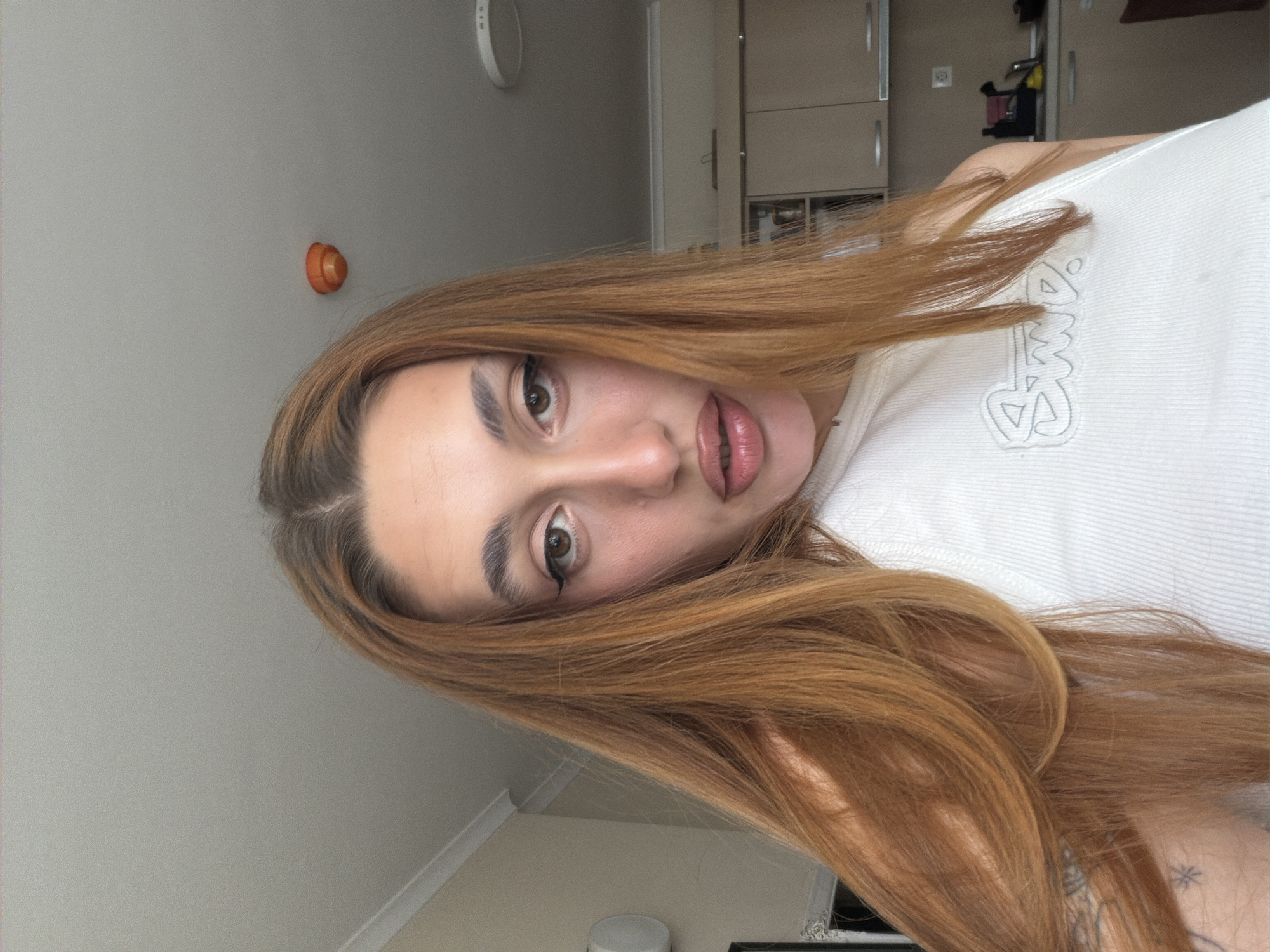 A young adult female, with long, reddish-brown hair parted on the left, is captured in a close-up self-portrait within an indoor residential setting in Sofia, Bulgaria. She is looking directly into the camera, her lips slightly parted and glossy. She wears dark eyeliner and has well-defined eyebrows. Her left arm, partially visible on the lower left, displays a small, star-shaped tattoo. She is wearing a white, ribbed tank top with a light grey embroidered design on the chest that appears to read "SMD."
The background features a light-colored ceiling with a white decorative molding along the wall junction on the upper left. An orange smoke detector is mounted on the ceiling in the upper center-right, and a circular ceiling light fixture is visible further to the right. To the immediate left, a white, circular wall-mounted object (possibly an air vent or speaker) is present. The mid-to-lower right background reveals a kitchen area with light-colored, possibly beige or cream, upper cabinets. A portion of a kitchen counter, sink, faucet, and a yellow sponge are partially visible beneath the cabinets.