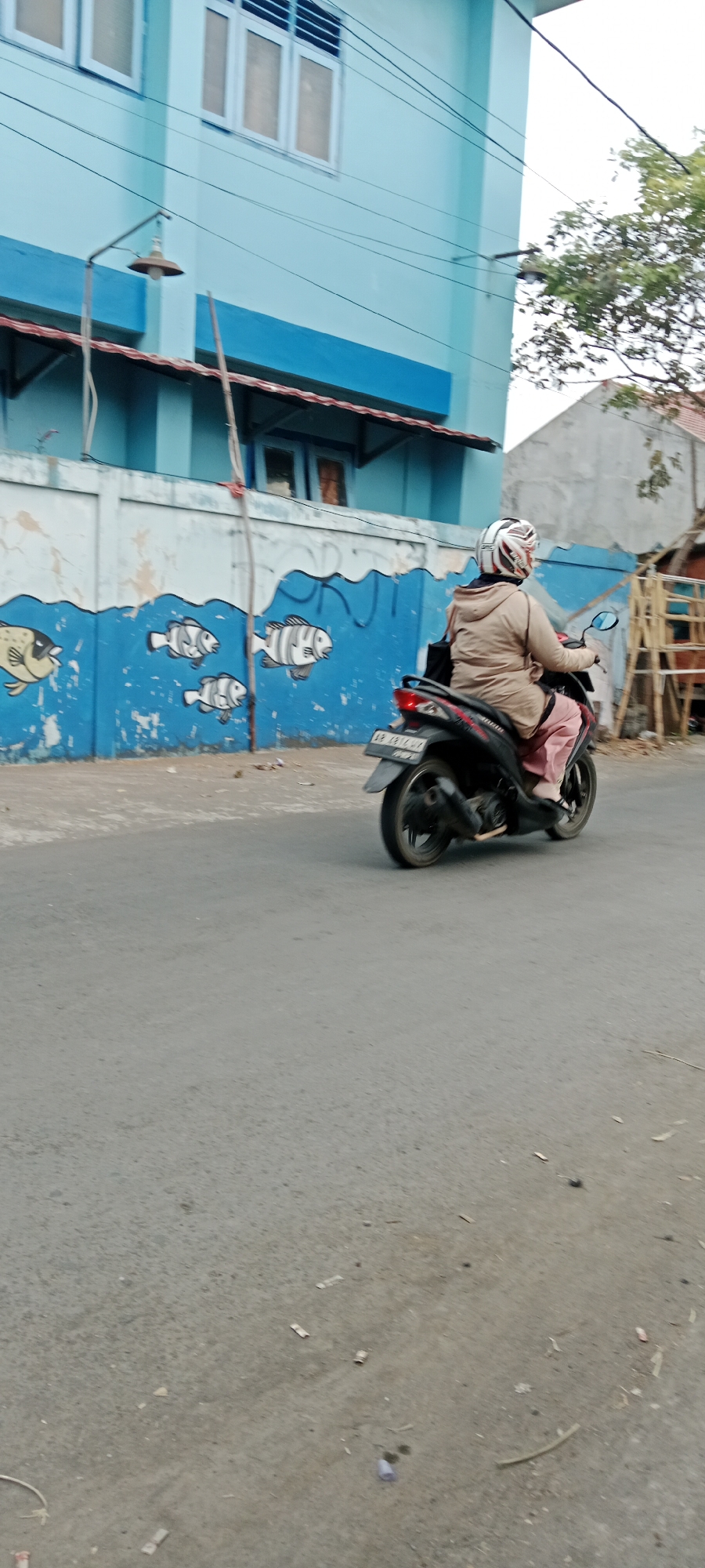 A person is riding a dark-colored scooter on a paved road. The rider is wearing a light-colored hooded jacket and light-colored pants, and a white and red helmet. The scooter has a license plate that reads "AB 2816 UY". In the background, there is a light blue building with windows and a mural of fish on a white wall. The mural depicts several fish swimming in blue water. To the right of the building, there is a stack of wooden pallets. The scene appears to be in an outdoor urban or semi-urban environment. The lighting suggests it is daytime, with overcast conditions. The road surface shows some debris and has a rough texture.