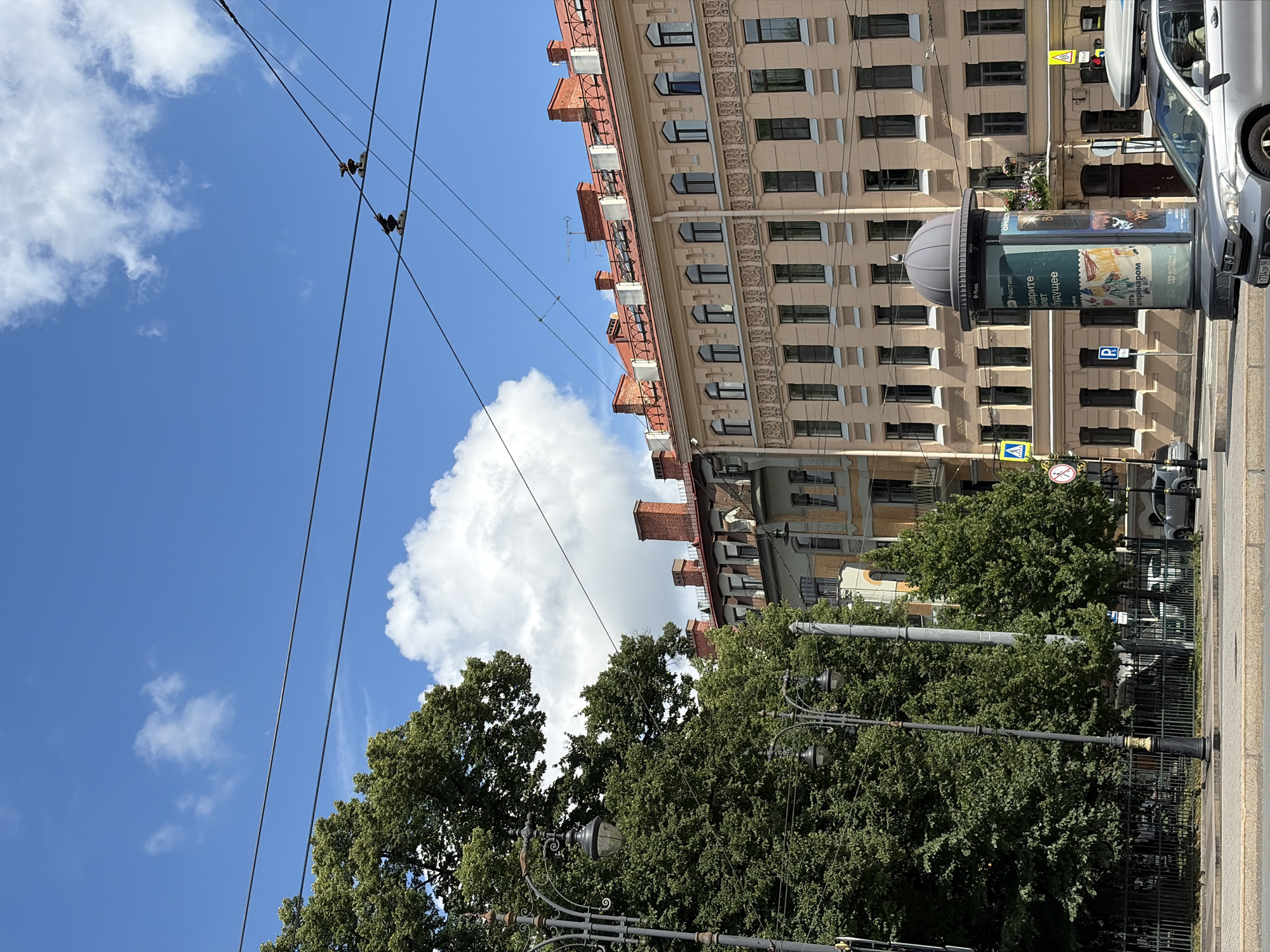 The image depicts a view of a city street in Saint Petersburg, Russia, on a sunny day with scattered clouds. In the foreground, a street with a sidewalk is visible. A silver car with a roof rack is parked on the right side of the street. To the left, there is a black metal fence and a tree-lined area with a decorative lamppost. Behind the fence, the tops of cars are visible.
The middle ground features several multi-story buildings. The most prominent building is a light peach-colored structure with a red tiled roof and numerous windows. Decorative elements are visible on the facade between the rows of windows. To the left of this building, another building with a gray facade and a red tiled roof is partially visible. In front of the peach-colored building, there is a cylindrical advertising pillar with posters on it.
The background consists of a bright blue sky with white clouds. Overhead, power lines stretch across the frame, supported by poles. Two small dark figures, likely birds, are perched on the power lines. The overall scene captures an urban environment with architectural elements characteristic of Saint Petersburg.