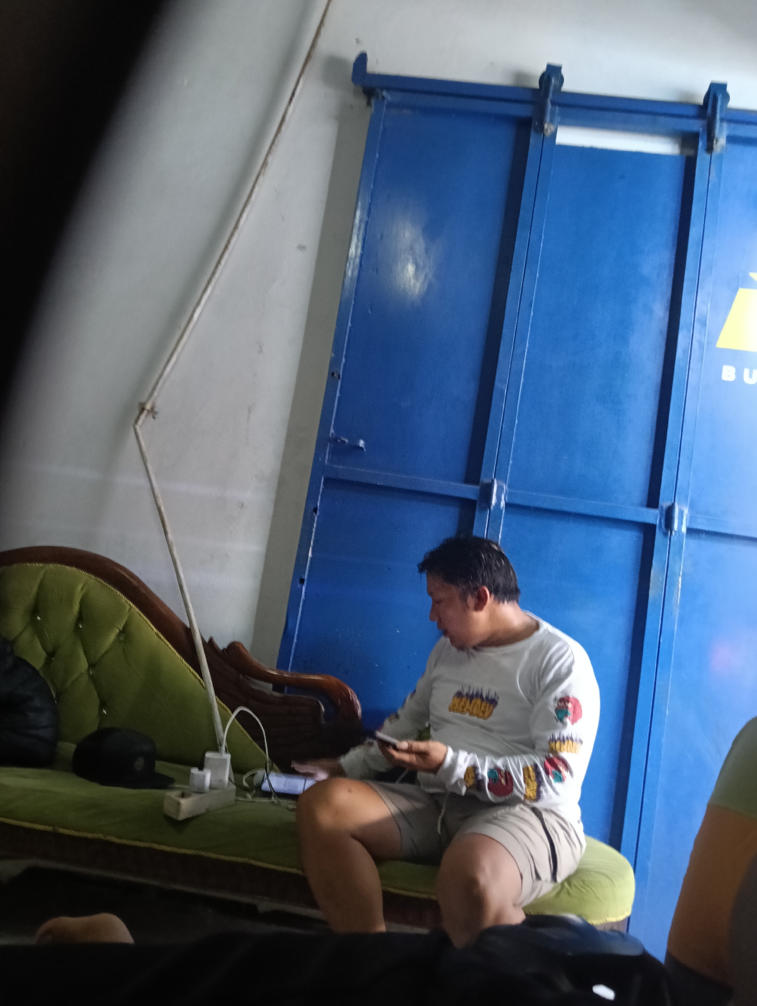 A young man sits on a green velvet couch, his left hand holding a mobile phone and his right hand resting on his lap. He is wearing a long-sleeved white shirt with colorful graphics on the sleeves and chest, and light brown shorts. The couch is situated in front of a large blue sliding door. On the couch next to the man is a black baseball cap and a power strip with several chargers plugged in. A thin, angled metal pole extends from the floor towards the ceiling on the left side of the frame, and a light is attached to its top. The room has white walls. The scene appears to be an indoor setting in Desa Cipakat, Indonesia.