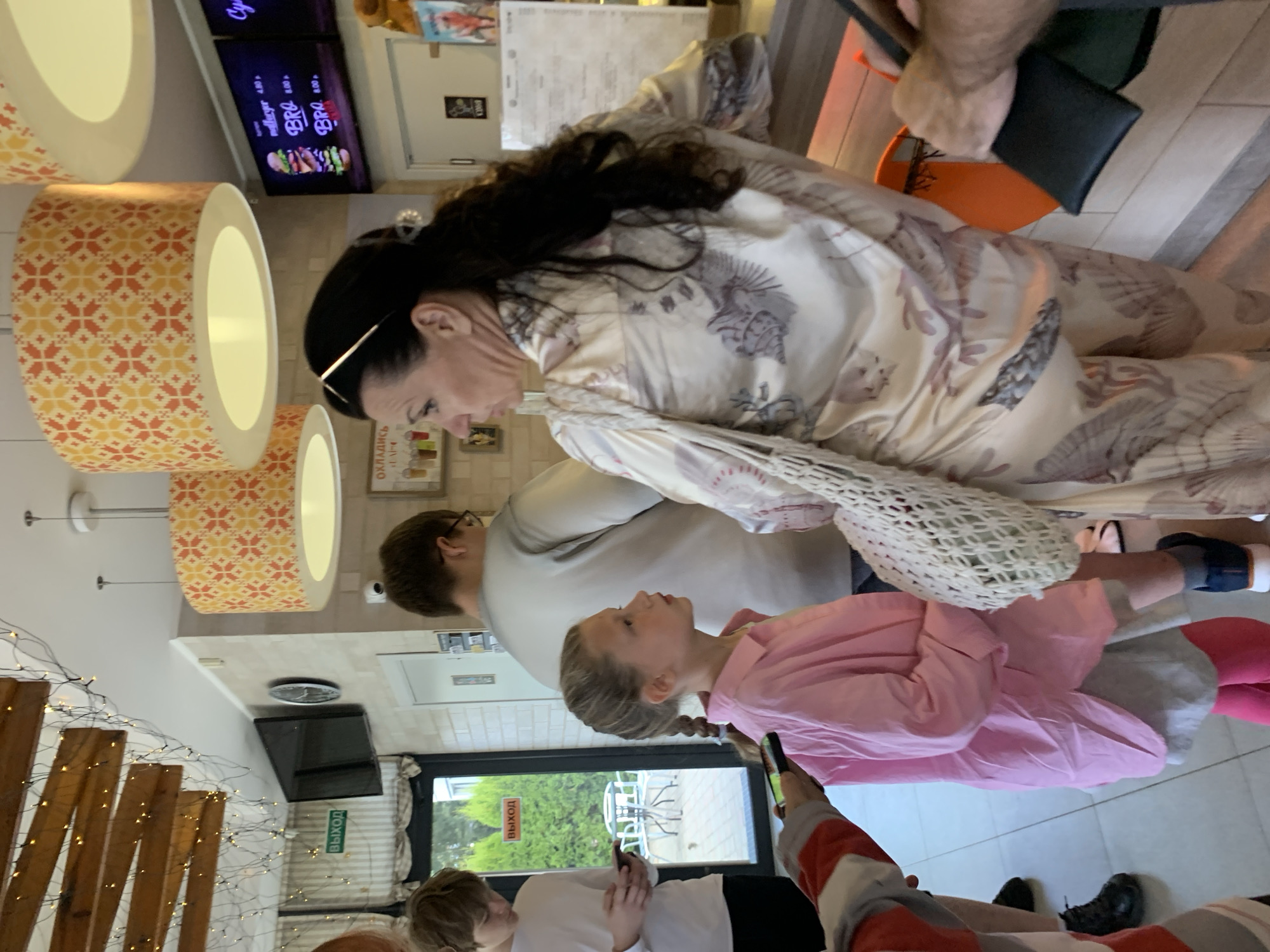 The image depicts a group of people standing indoors, possibly a cafe or fast-food establishment, in Sluck, Belarus. In the foreground, a young girl with braided hair, wearing a pink button-up shirt and grey shorts over pink leggings, looks to her right. To her left, a woman with dark hair pulled back, wearing a light-colored patterned robe, faces the girl and appears to be speaking. She has a white crocheted bag slung over her shoulder. Behind the girl and woman, a young man in a white t-shirt holds a smartphone, looking down. Further in the background, another person in a light grey shirt is partially visible. The environment includes ceiling lights with patterned shades, a television screen displaying a menu with burger images, and a sign with Cyrillic text. A door with a green "EXIT" sign is visible on the left. The overall lighting suggests an indoor setting with artificial illumination.