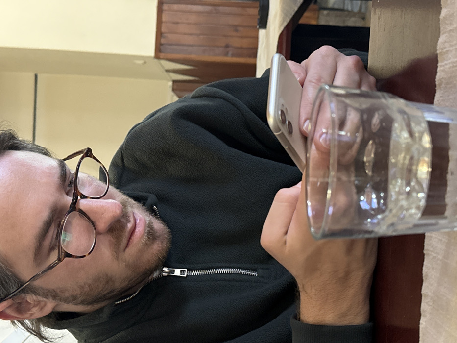 An individual, presumed male, is seated at a table in an indoor establishment within Tutin Municipality, Serbia. The subject wears a dark, possibly black or dark green, fleece half-zip pullover and tortoise-shell framed eyeglasses. The individual's gaze is directed downwards, focused intently on a light-colored smartphone held horizontally with both hands. The smartphone's rear is visible, featuring two camera lenses. In the immediate foreground, a clear cylindrical glass, approximately half-filled with clear liquid, rests on a light-colored cloth or placemat, which is atop a dark brown table surface. The background wall is bifurcated, displaying a pale yellow upper section and a lower section clad with dark brown wooden paneling. A portion of another table covered with a light-colored textile is visible in the distant right background.