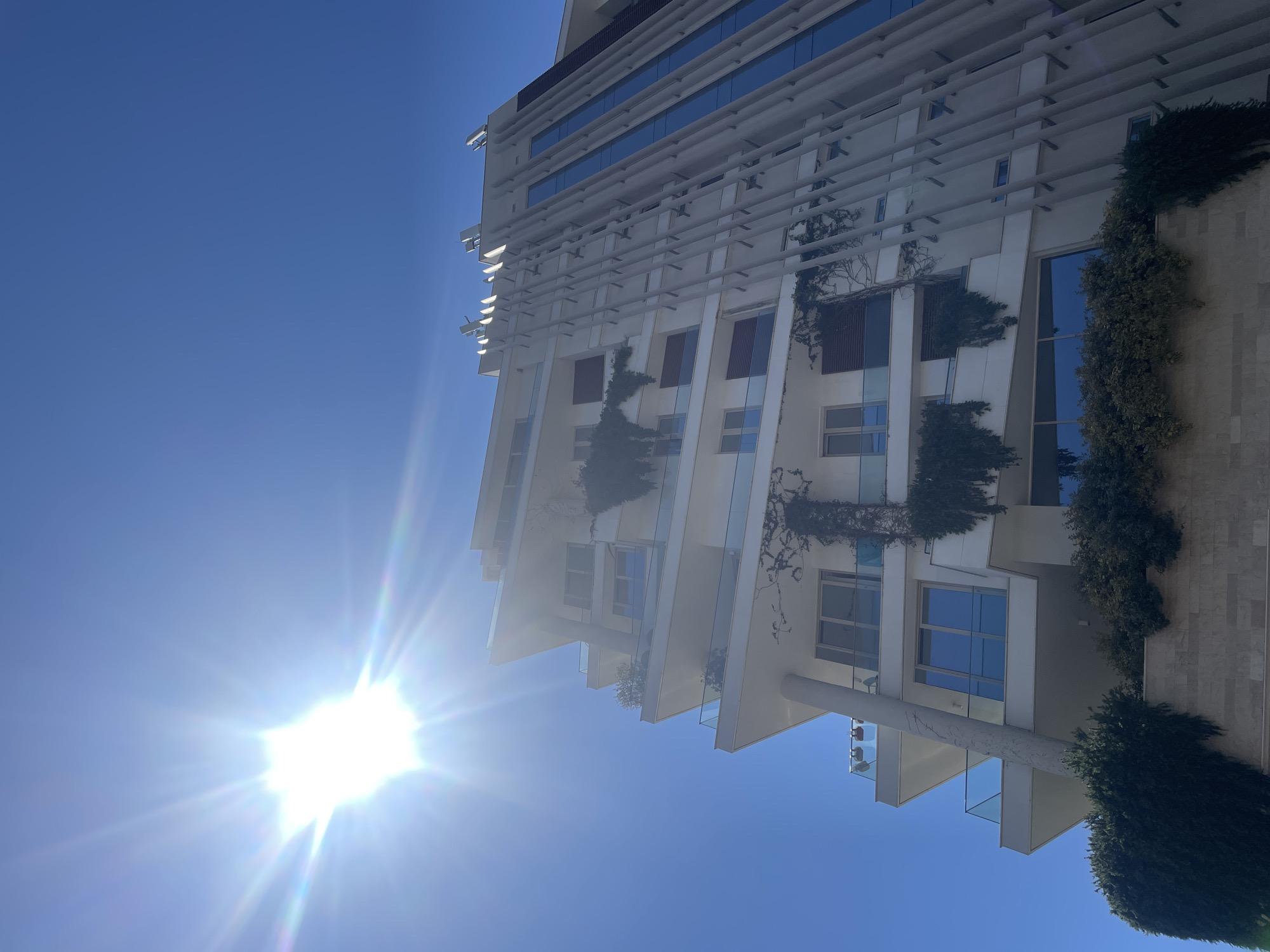 In a low-angle shot, a modern, multi-story white building with staggered balconies and large windows dominates the frame against a clear blue sky. The sun shines intensely in the upper left quadrant, creating a lens flare that streaks across the sky and illuminates the building. Green vines and plants are draped over several balconies and along the side of the building, adding a touch of nature to the architecture. The building features a unique facade on the right side, with vertical, light-colored panels and a tall, blue-tinted glass section. The foreground shows a stone-like base with dense green foliage. The scene depicts an urban setting with striking contemporary architecture bathed in bright sunlight.
