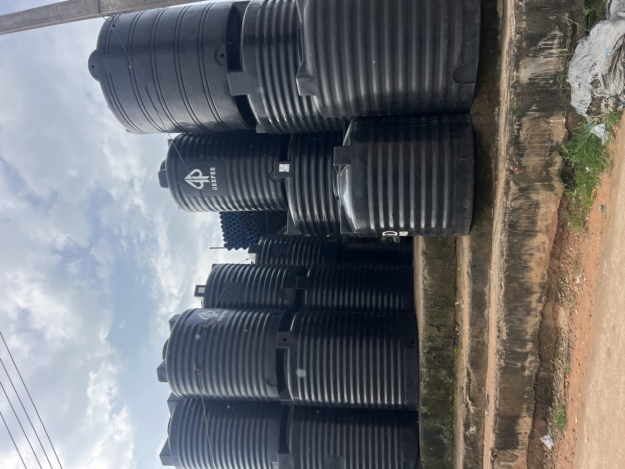 The image shows a street scene in Okpanam, Nigeria, featuring a large stack of black plastic water tanks. The tanks are of various sizes and are arranged in a tiered formation on a concrete platform. Some of the tanks display the "GEEPEE" logo. In the background, a stack of blue pipes and a utility pole are visible, along with a cloudy sky. The ground in front of the platform is a mix of dirt and debris, with some sparse vegetation. The scene appears to be an outdoor storage or sales area for water tanks, likely on a commercial street.