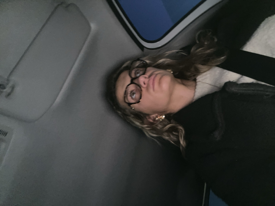 Inside a car, a young woman with glasses and blonde hair sits angled towards the viewer. She wears a dark coat and a lighter-colored top underneath. A seatbelt is visible across her chest. To her left is a car window showing a glimpse of the sky and a sun visor. The interior of the car is predominantly gray. The woman's expression is slightly upward-looking, and her lips are pursed.