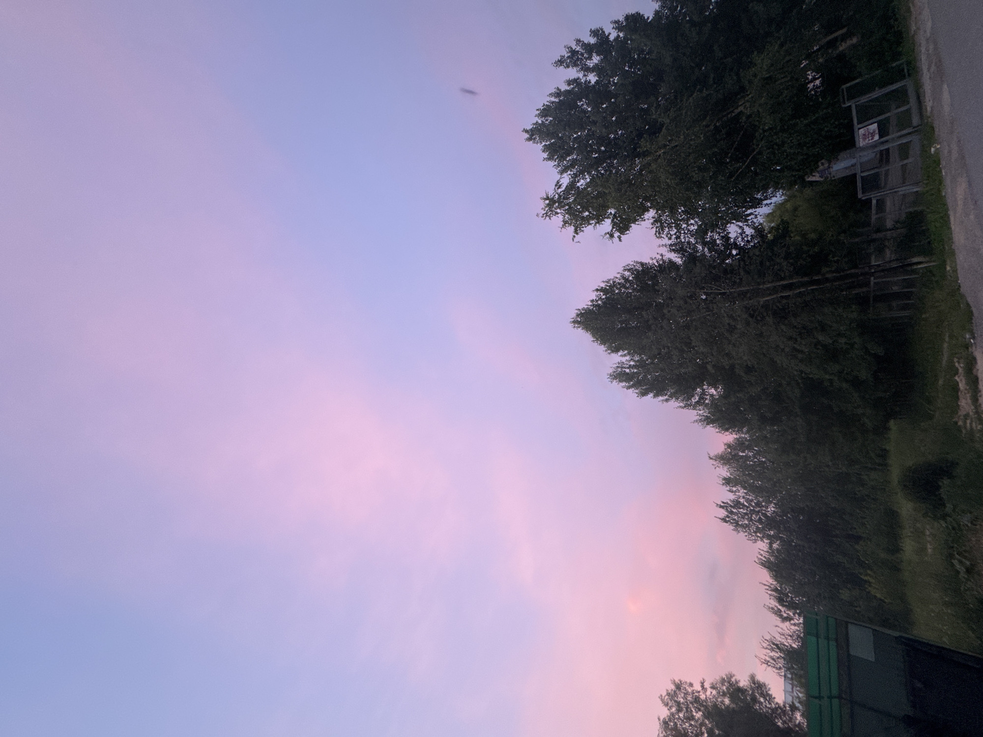The image depicts a scenic twilight sky with vibrant pink and purple hues above a line of trees and vegetation. In the foreground, a dark, angular structure, possibly a fence or a section of a building, is visible on the left. A paved surface, likely a road, is present at the bottom right of the image. There are no people or animals visible. The overall scene suggests an outdoor environment, potentially on the outskirts of a settlement, with the characteristic colors of a sunset or sunrise dominating the sky. The location is identified as the city of Megion, Russia.