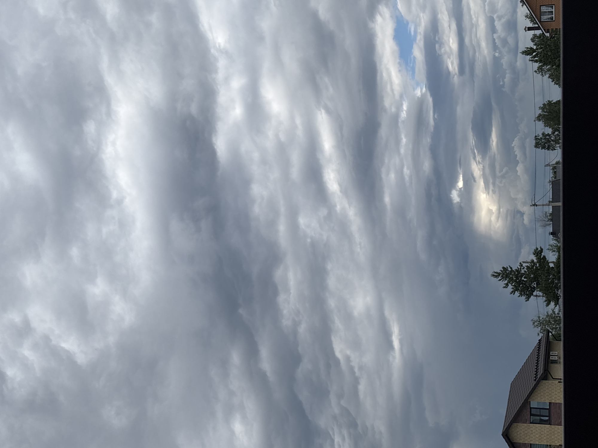 The image captures a dramatic sky filled with a dense pattern of undulating, gray clouds. These clouds stretch across the frame, appearing textured and layered, with lighter, brighter patches suggesting the presence of the sun attempting to break through. In the upper right portion of the image, a sliver of clear blue sky is visible, providing a stark contrast to the overcast conditions.
Along the right edge of the frame, the scene transitions to the terrestrial realm, revealing the upper floors of a two-story house with a dark-tiled roof and a few windows. Adjacent to the house are the tops of several trees, their foliage appearing dense and green. A power line is also visible, running horizontally across the scene. The overall impression is that of a temperate, possibly overcast day in a residential area, with the sky dominating the view. The heavy cloud cover and the way the light interacts with them suggest a dynamic weather system, possibly indicating approaching rain or a recent storm. The location is identified as Alexandrovka, Russia. No people or text are visible in the image.