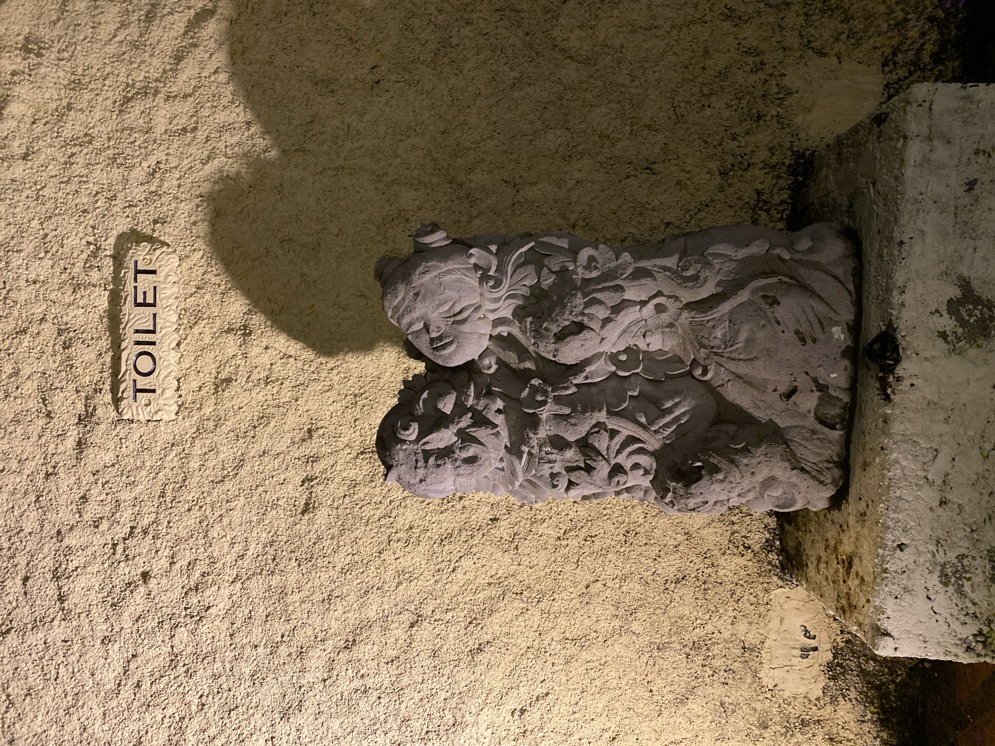A stone sculpture of two figures is positioned on a stone pedestal in front of a textured, light-colored wall. Above the sculpture, a sign is affixed to the wall, reading "TOILET". The wall is illuminated by artificial light, casting shadows from the sculpture onto it. The overall scene appears to be an outdoor setting, likely a public restroom area. The image does not contain any individuals or ongoing actions. The location is identified as Kuta Selatan, Indonesia.